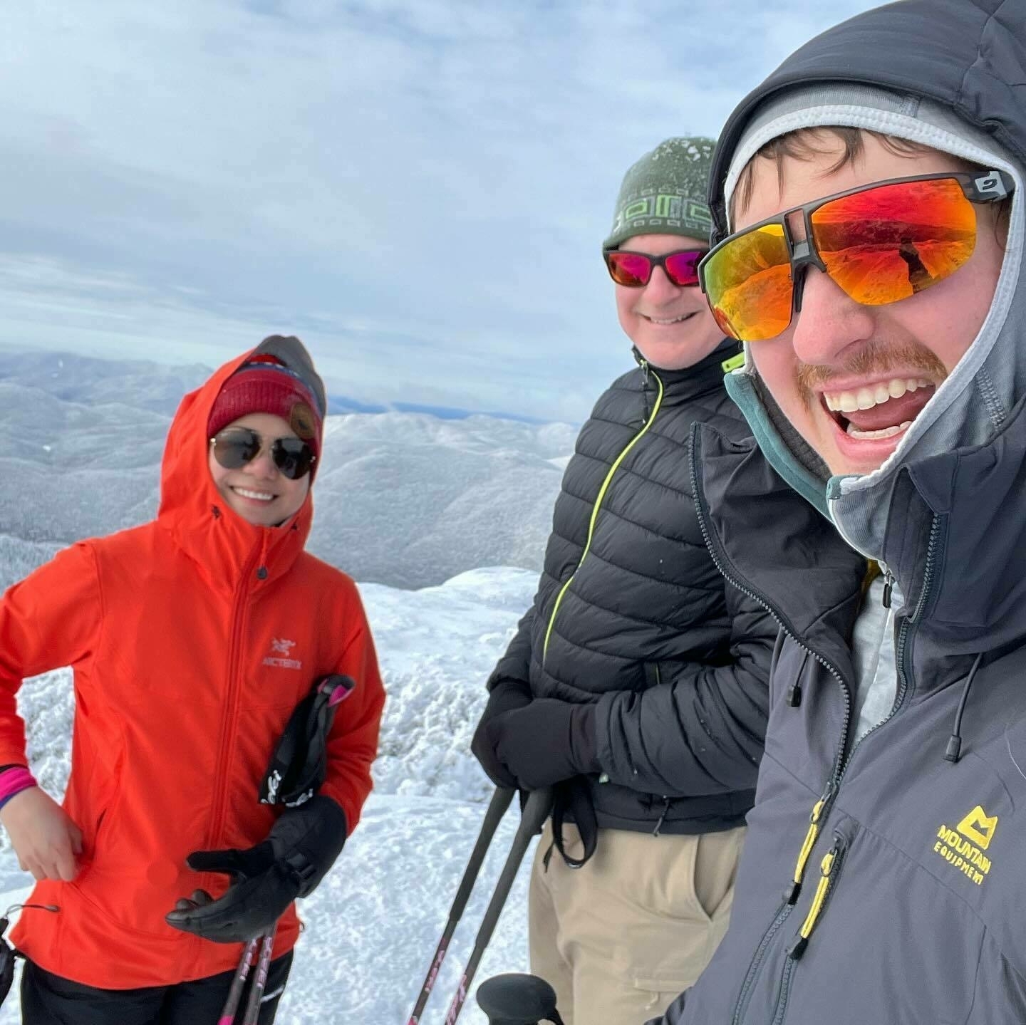 Three individuals standing atop a snowy mountain, smiling for a selfie, surrounded by a snow-covered landscape under a cloudy sky.