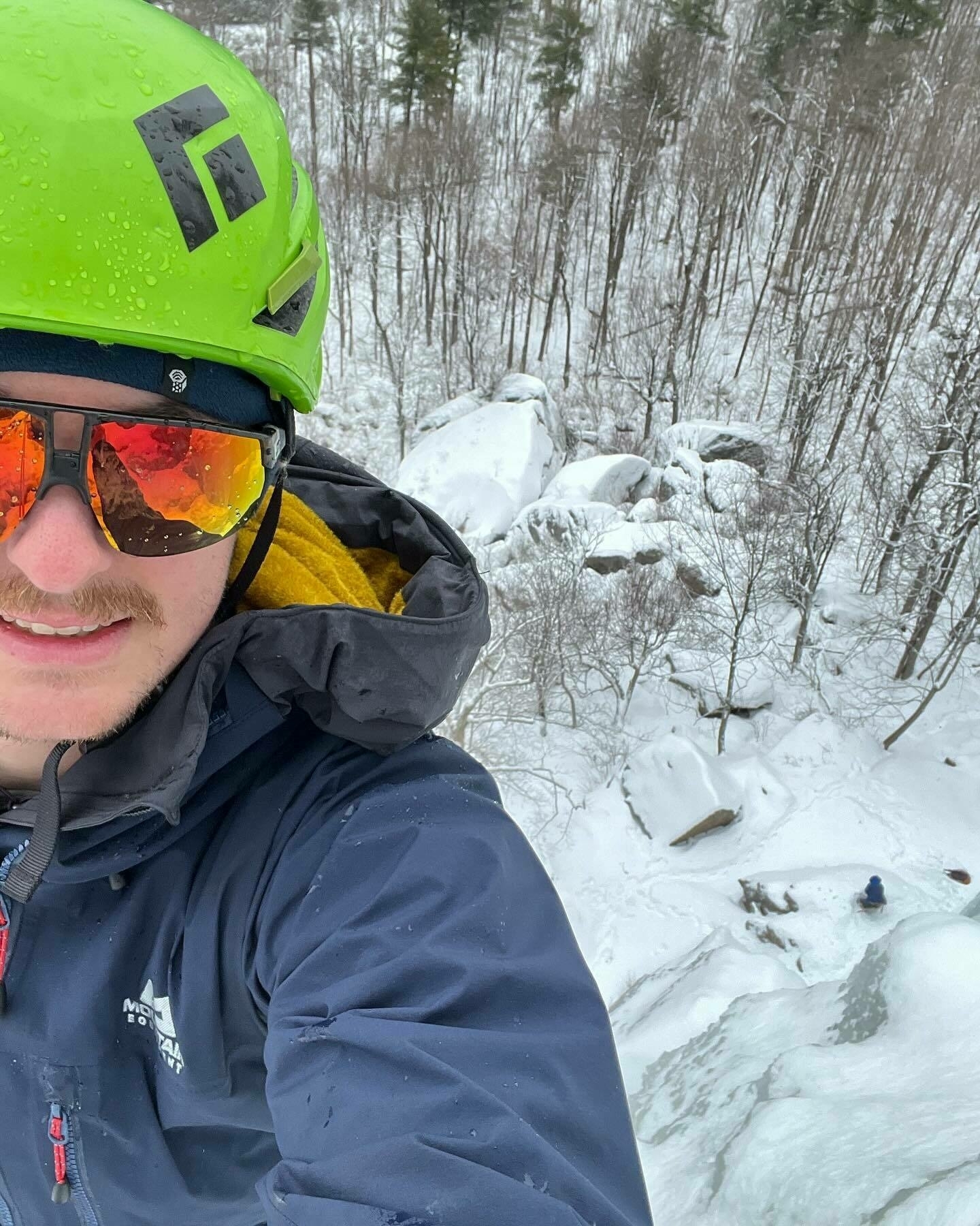 A person in a green helmet and sunglasses is taking a selfie in a snowy forested area with rocks partially covered in snow.