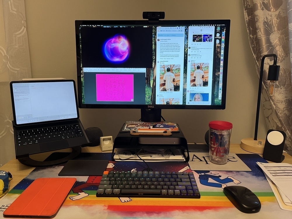 A computer monitor displaying various windows, a tablet on a stand, and a keyboard and mouse are on a desk with colorful decorations, surrounded by office supplies and beverages.