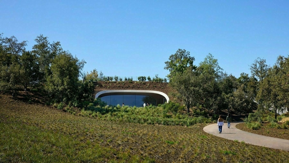 A modern, earth-sheltered building with a large, curved glass facade is nestled into a grassy hillside. Two people walk on a winding path surrounded by trees and greenery.