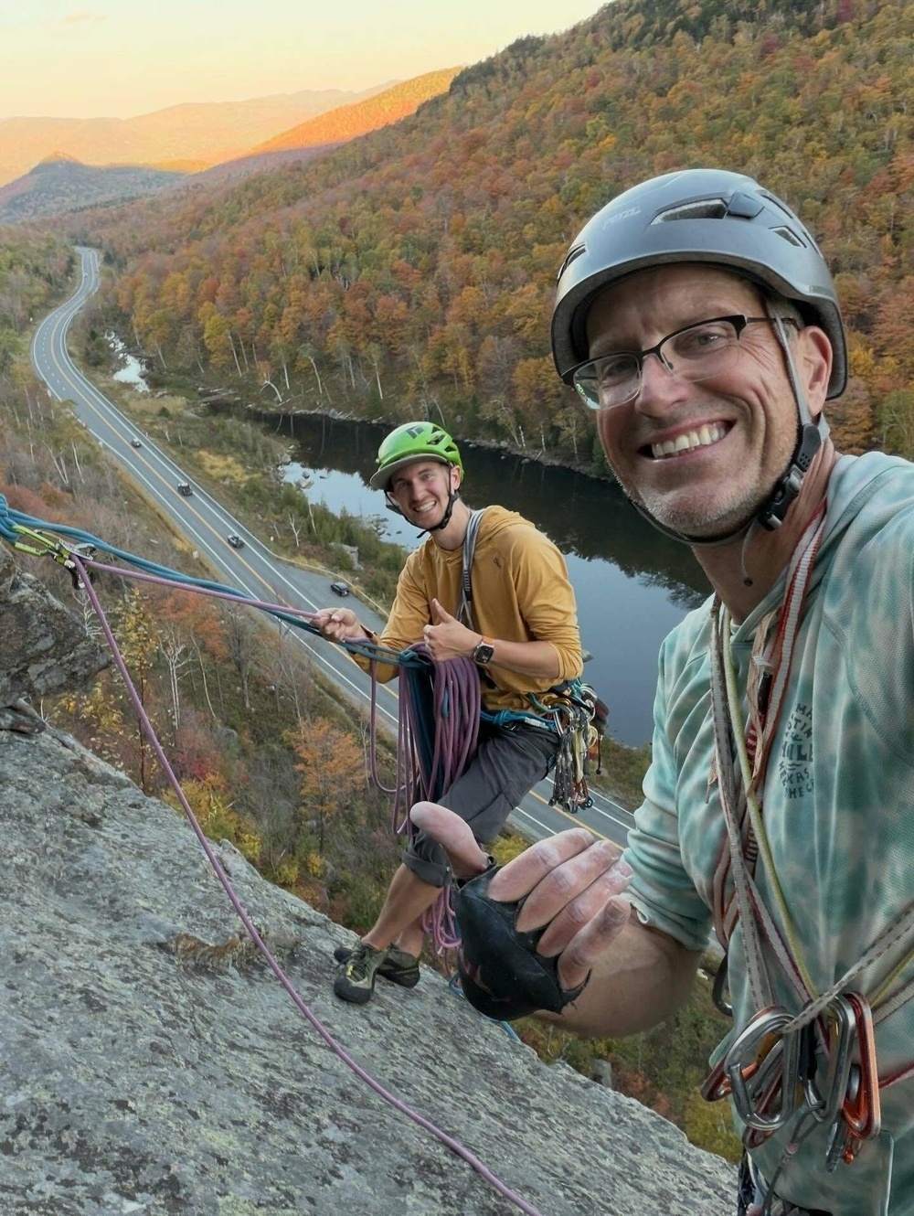 Two climbers wearing helmets and gear smile while scaling a rock face with ropes high above a winding road and river amidst an expanse of colorful autumn forested mountains.