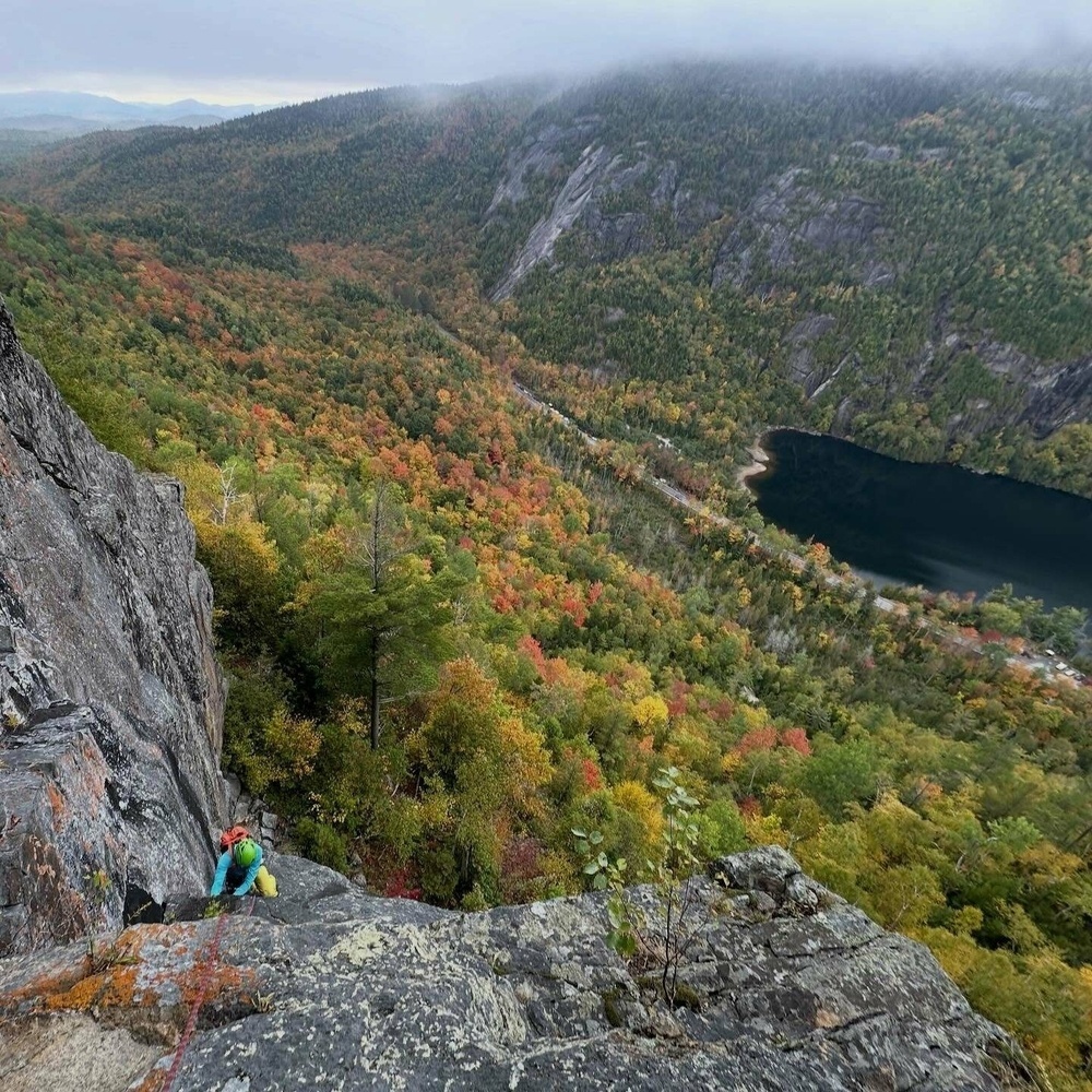 Climber ascending a large rocky cliff face surrounded by dense forest with autumn foliage and a nearby dark lake under a cloudy sky in a mountainous landscape.