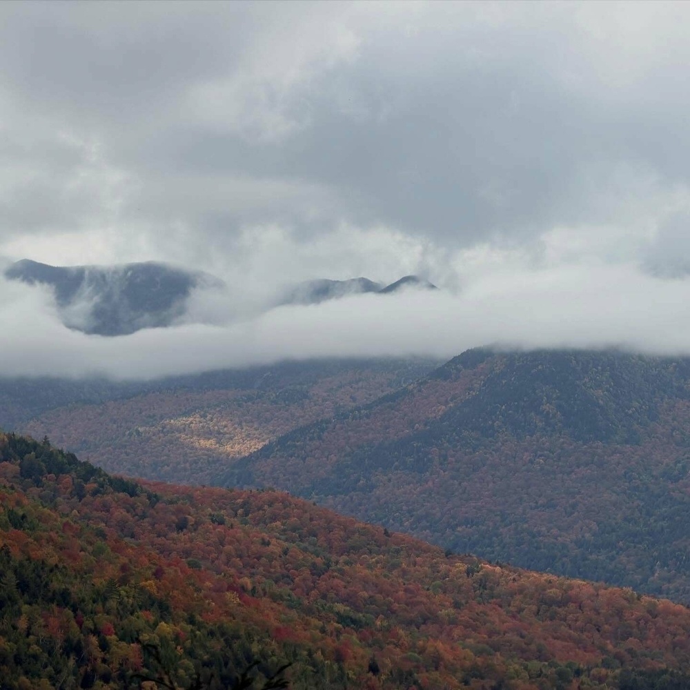 Clouds hover above mountain peaks covered in colorful autumn foliage, creating a serene, misty atmosphere in a vast, hilly landscape.