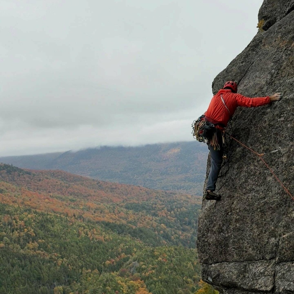 A climber in a red jacket scales a steep rock face surrounded by lush green and autumn-colored forested mountains under a cloudy sky.