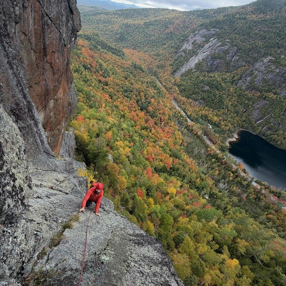 A climber in red ascends a steep rock face using a rope amidst a vibrant autumn forest with a river and lake visible below.