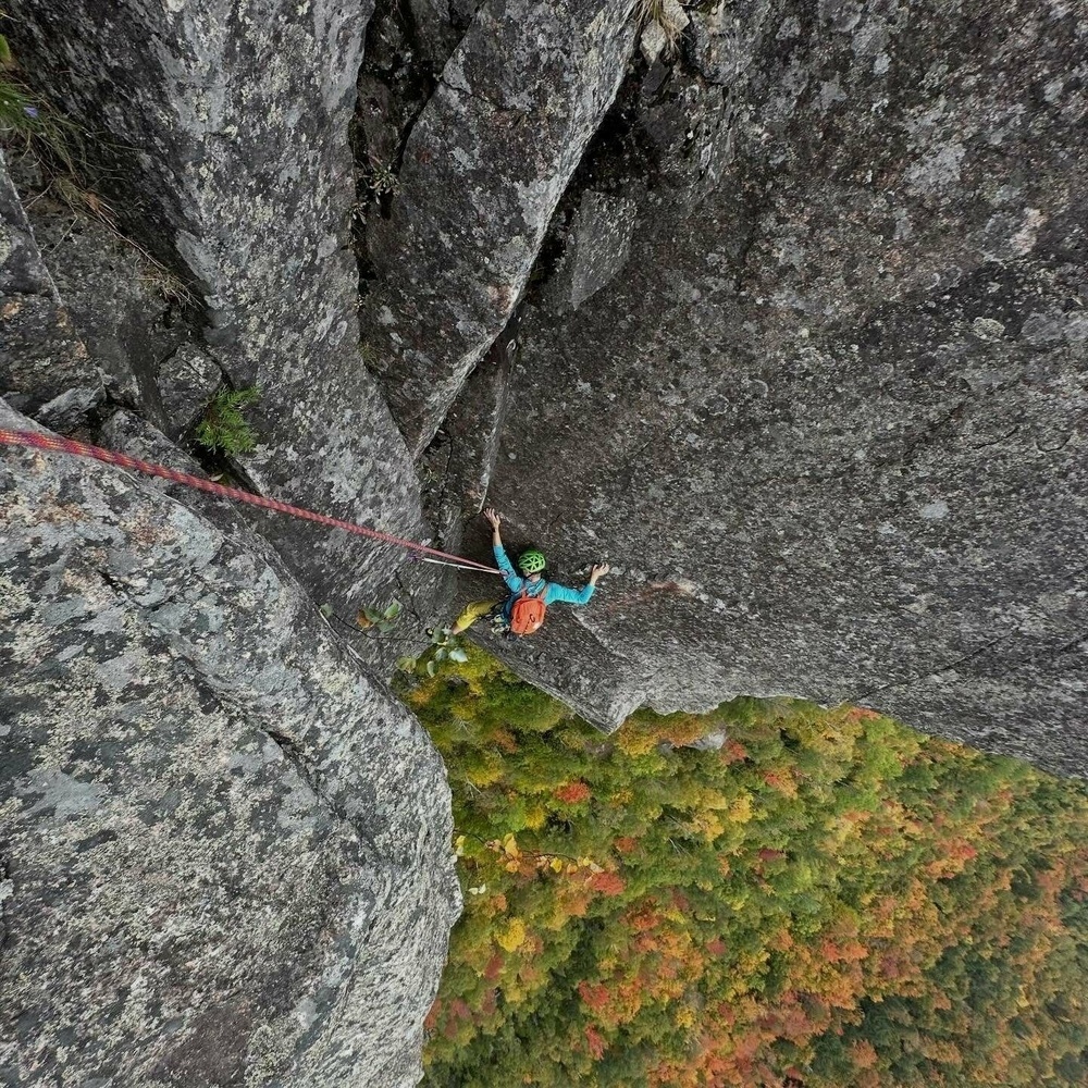A climber wearing a helmet and backpack ascends a steep rock face attached to a red rope overlooking a forested landscape with autumn foliage below.
