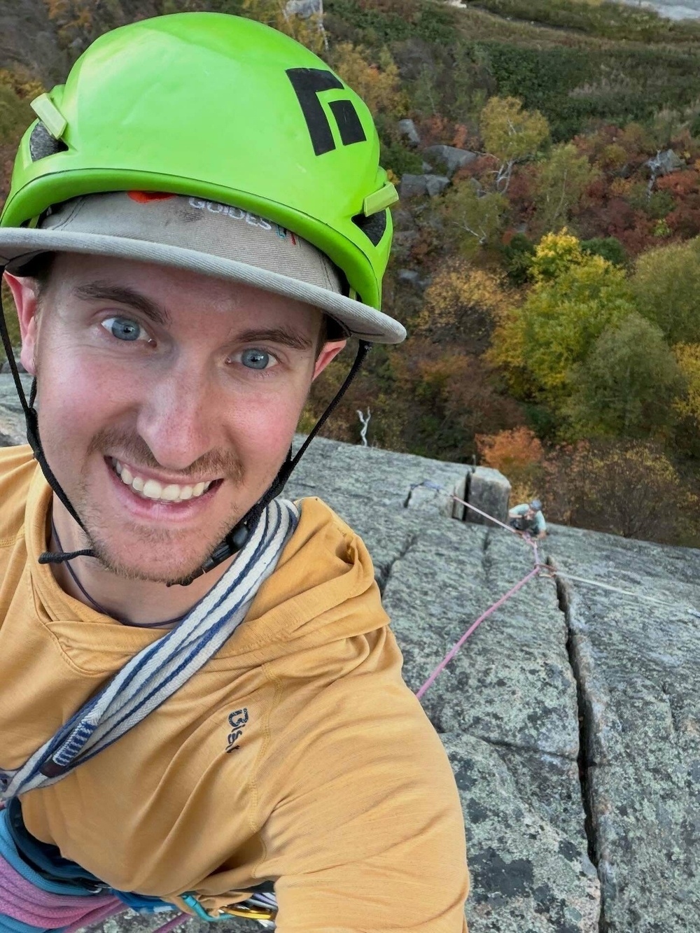 A person wearing a green helmet smiles at the camera while rock climbing. They are secured with ropes against a large rock face surrounded by colorful autumn foliage.