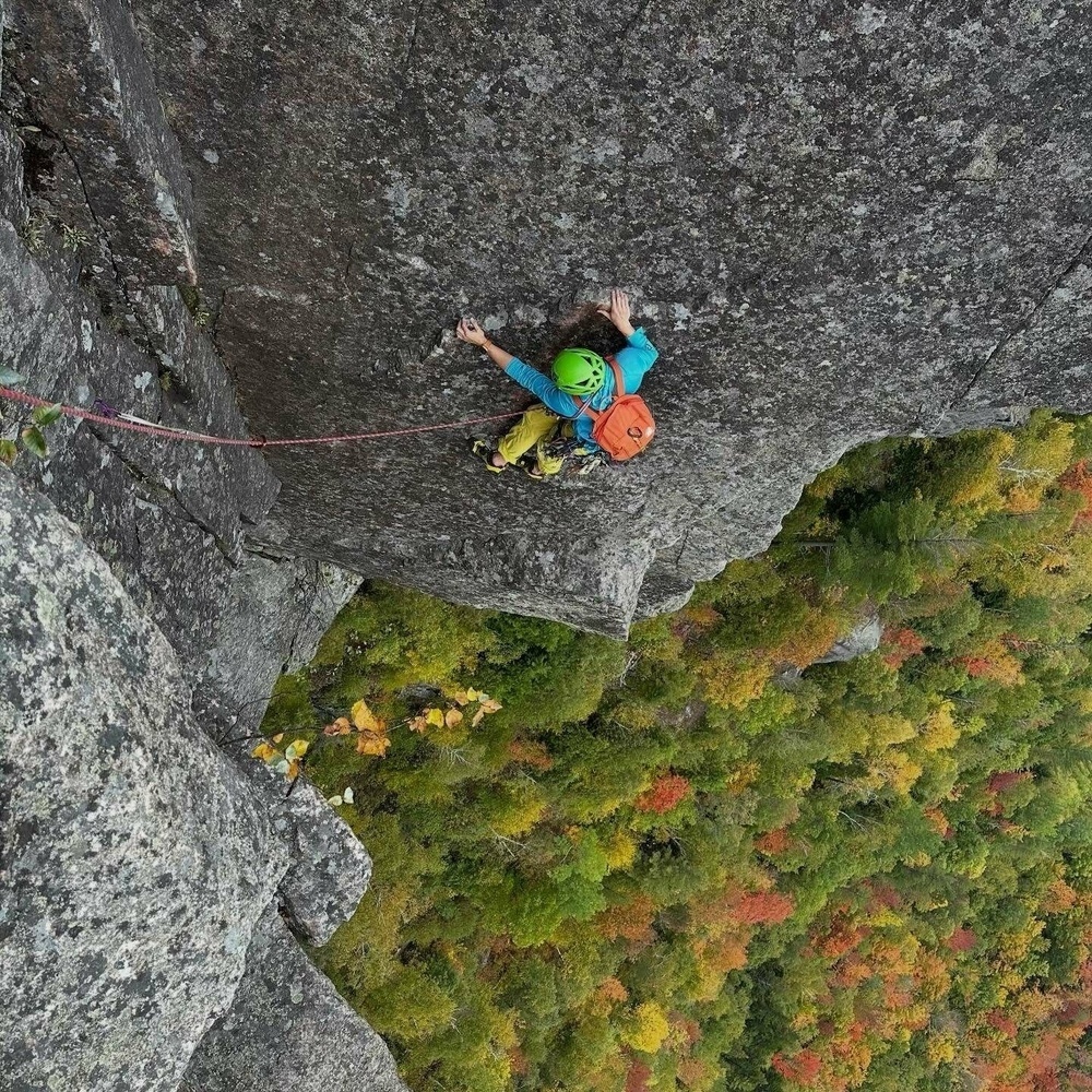 A person climbs a vertical rock face gripping tightly wearing safety equipment. An autumn forest with colorful foliage stretches below providing a vibrant scenic backdrop.