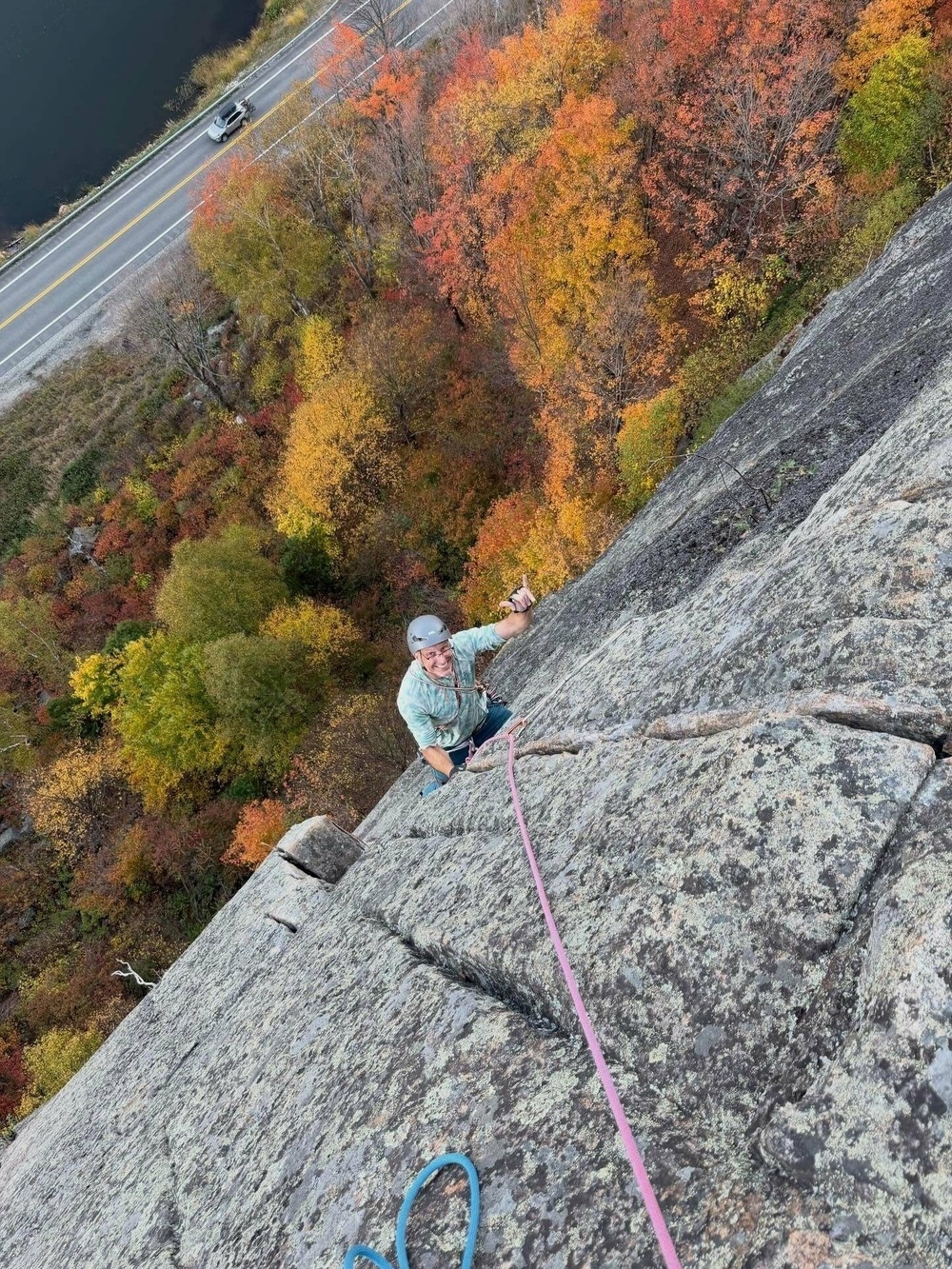 Climber ascends a steep, rocky cliff using a rope, amid vibrant autumn trees and overlooking a winding road with a car below.