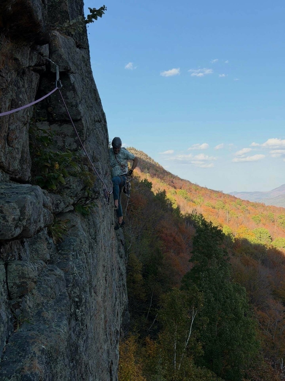 A climber ascends a steep rock face using climbing gear surrounded by a vast landscape of autumn-colored trees under a clear blue sky.