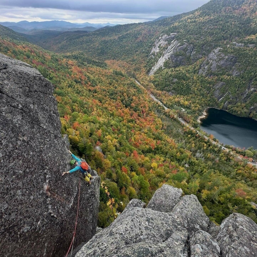 A climber scales a steep rocky cliff, surrounded by vibrant autumn foliage and overlooking a winding road and calm lake below.