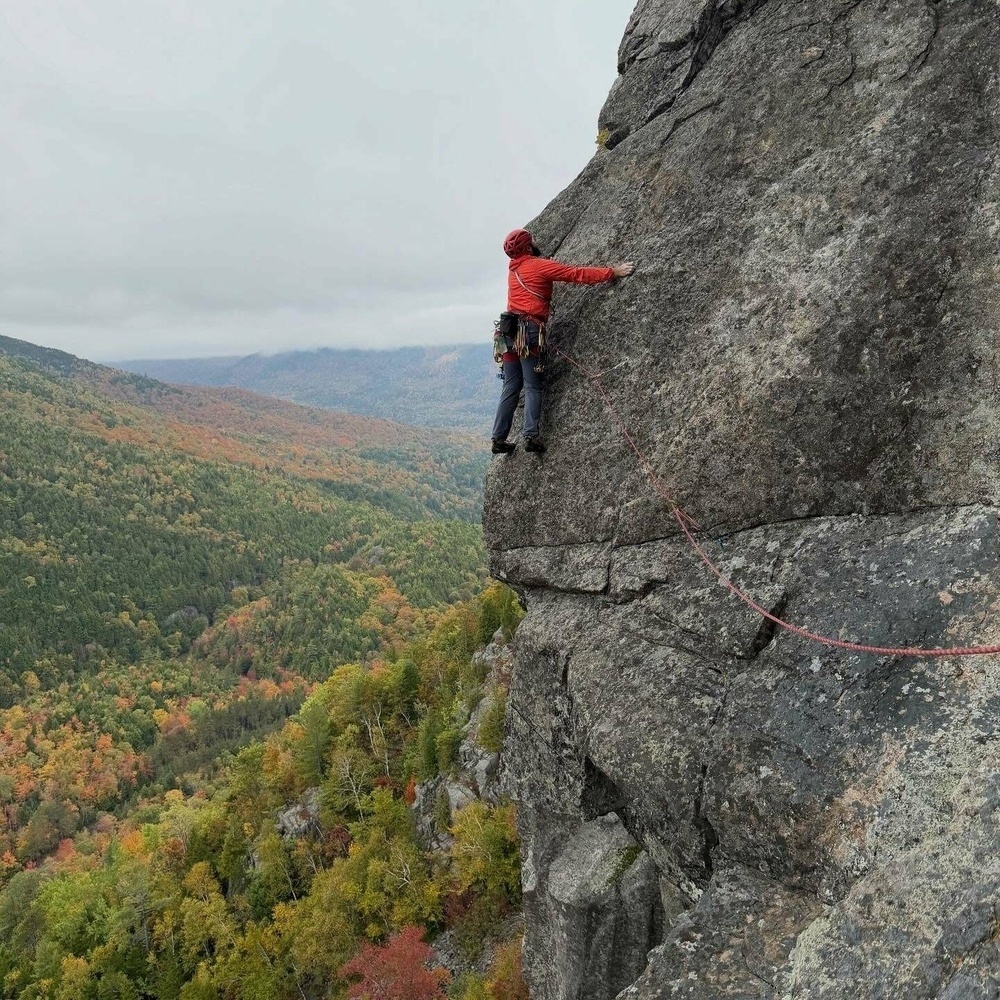 A climber ascends a steep rock face using safety gear and ropes against a backdrop of expansive, colorful autumn forests stretching to the horizon under an overcast sky.