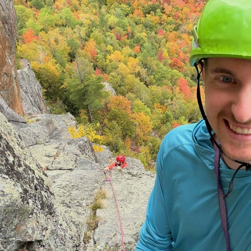 A climber wearing a green helmet smiles while another climber ascends a rocky cliff. Vibrant autumn foliage covers the surrounding forested landscape below.