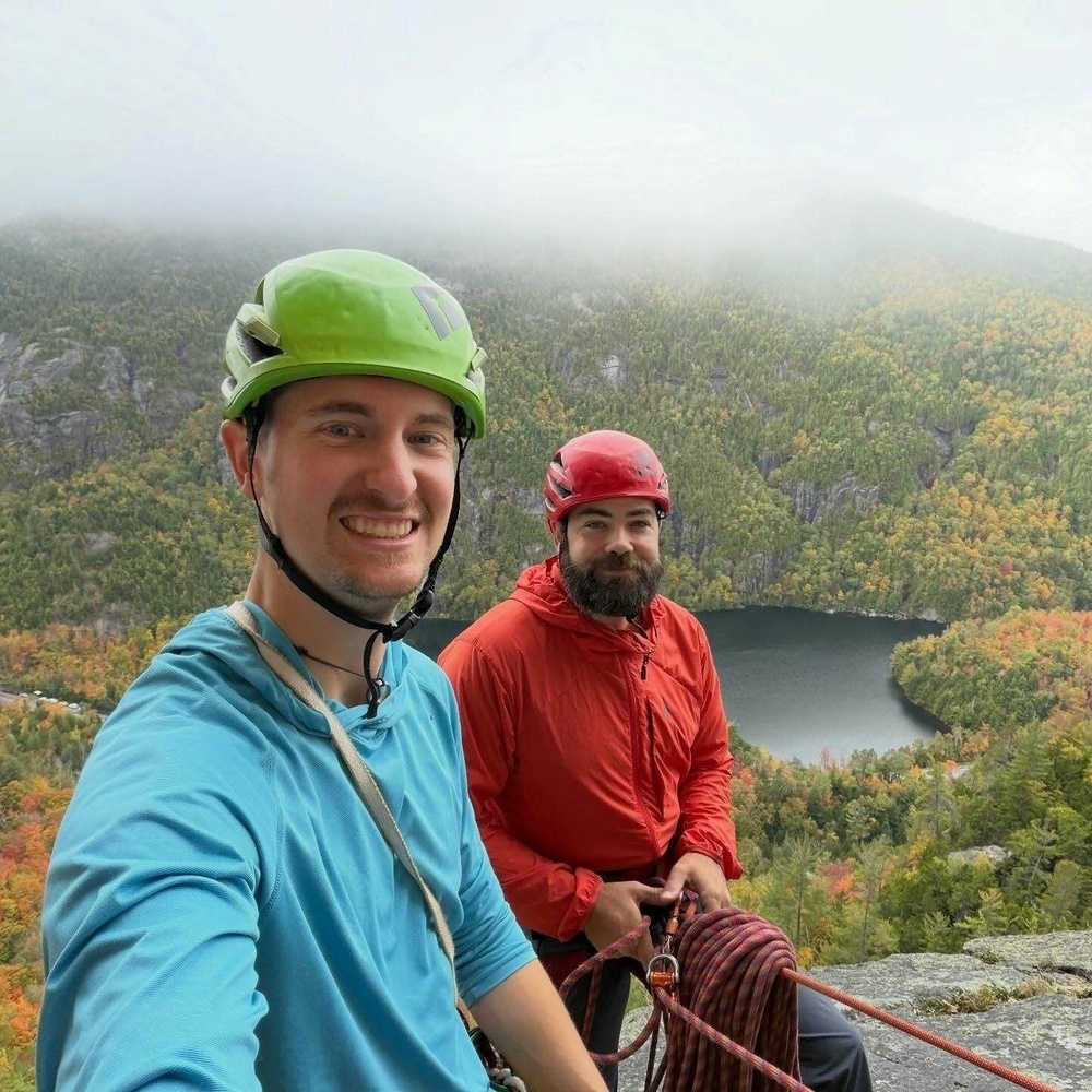 Two climbers wearing helmets smile atop a mountain holding climbing ropes overlooking a forested landscape and a lake partially obscured by clouds