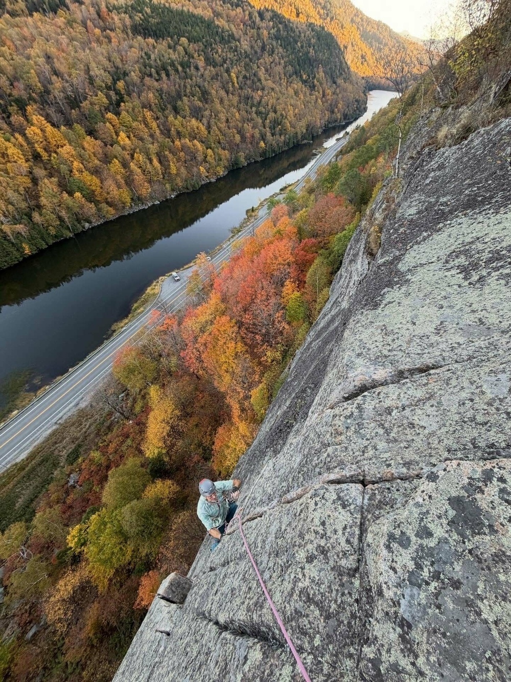 Climber ascending steep rock face secured by a rope surrounded by vibrant autumn foliage with a river and road in the valley below.