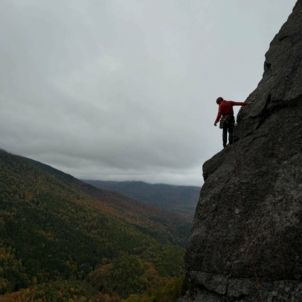 Climber ascends steep rock face under overcast sky with forested mountains in the background.