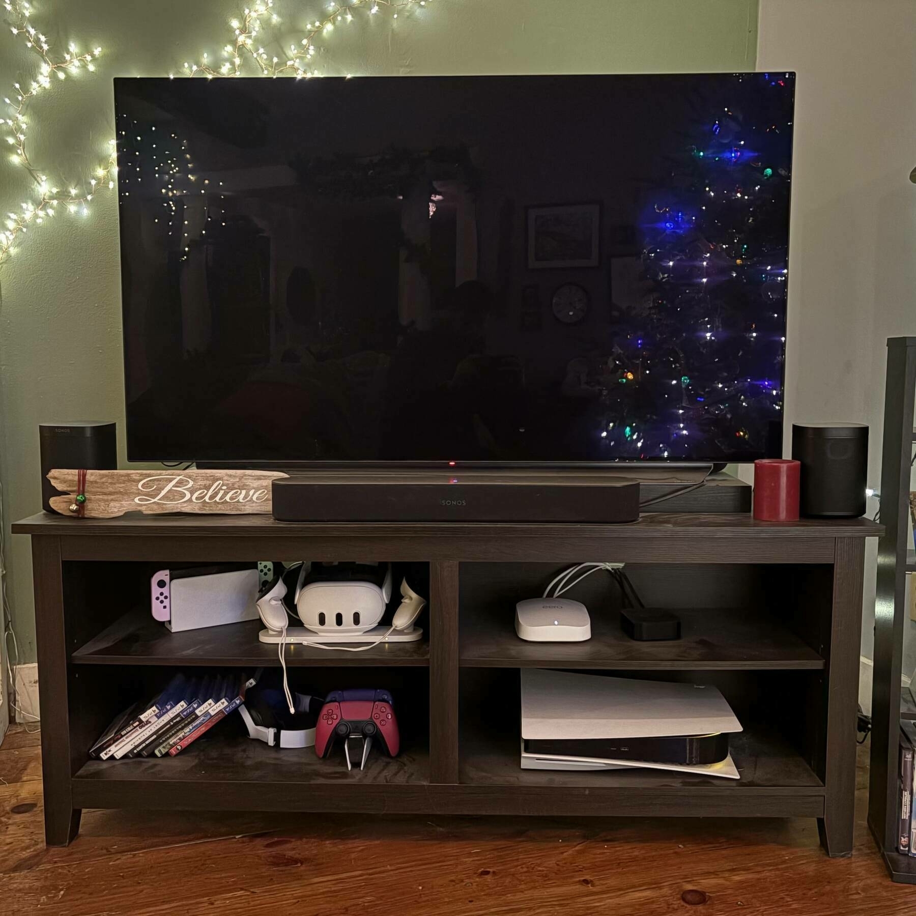 Auto-generated description: A television is set up on a stand with gaming consoles, controllers, and small speakers, next to a decorated Christmas tree with string lights on the wall.