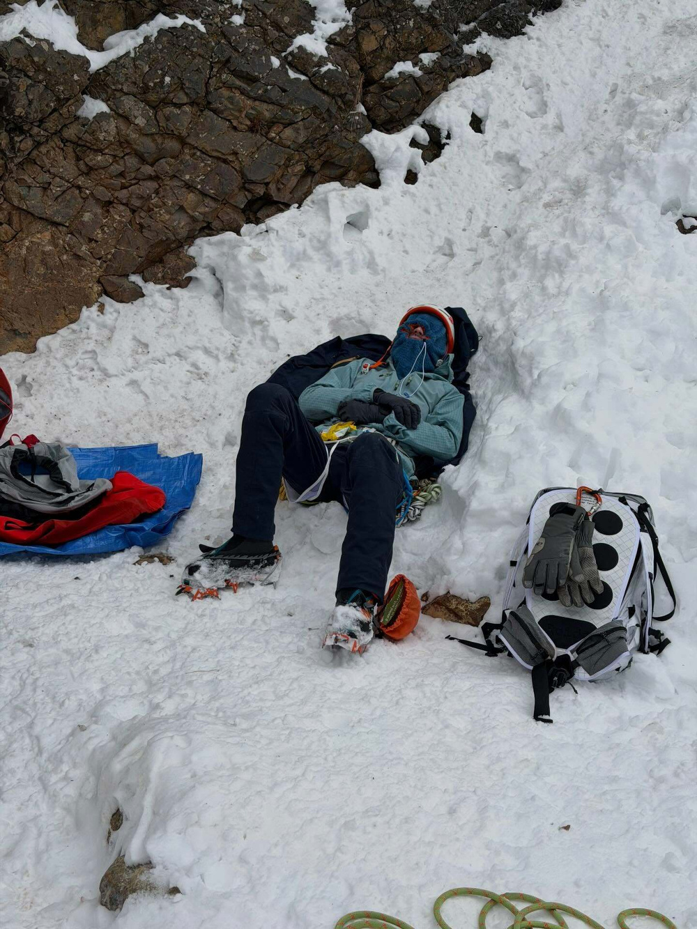 A climber rests on snowy ground, wearing crampons and bundled in warm gear. Nearby, a backpack, gloves, and climbing ropes are set against a rocky snowy backdrop.