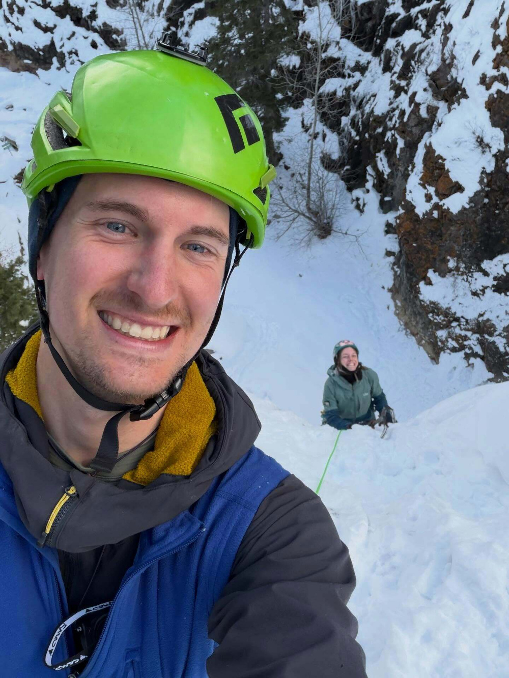 A person wearing a green helmet smiling in the foreground while another person climbs a snow-covered slope using a rope in a snowy mountainous area.