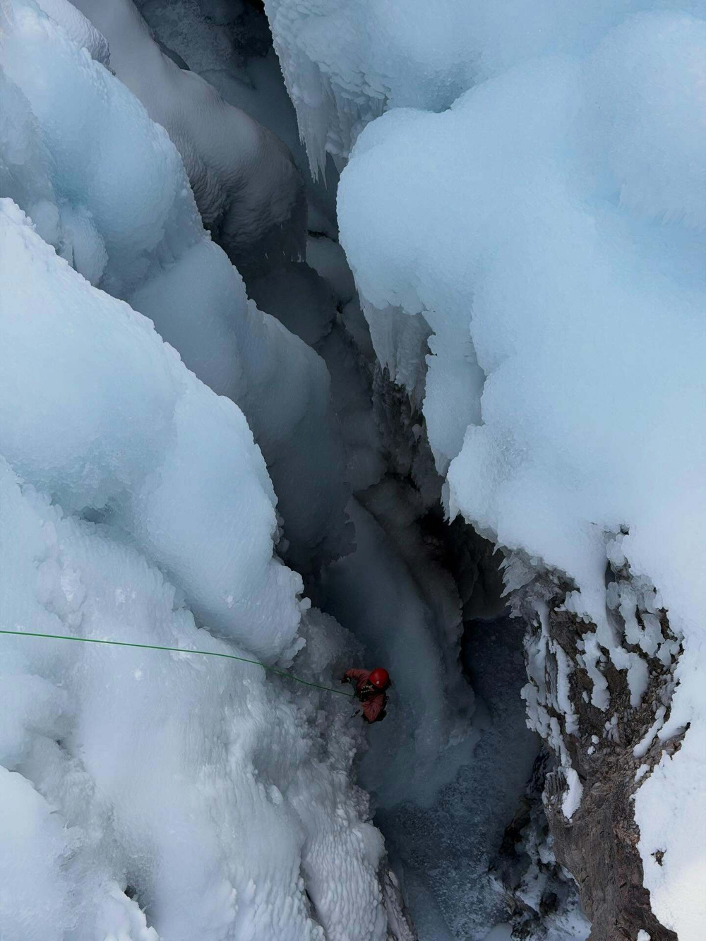 A climber ascends an icy crevasse using a rope surrounded by rugged ice formations and snowy surfaces in a mountainous environment.
