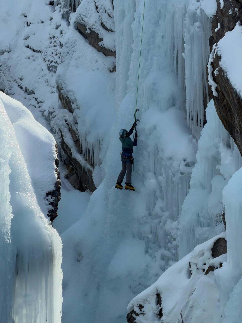 Climber ascending a frozen waterfall using an ice axe in a snowy mountainous landscape.