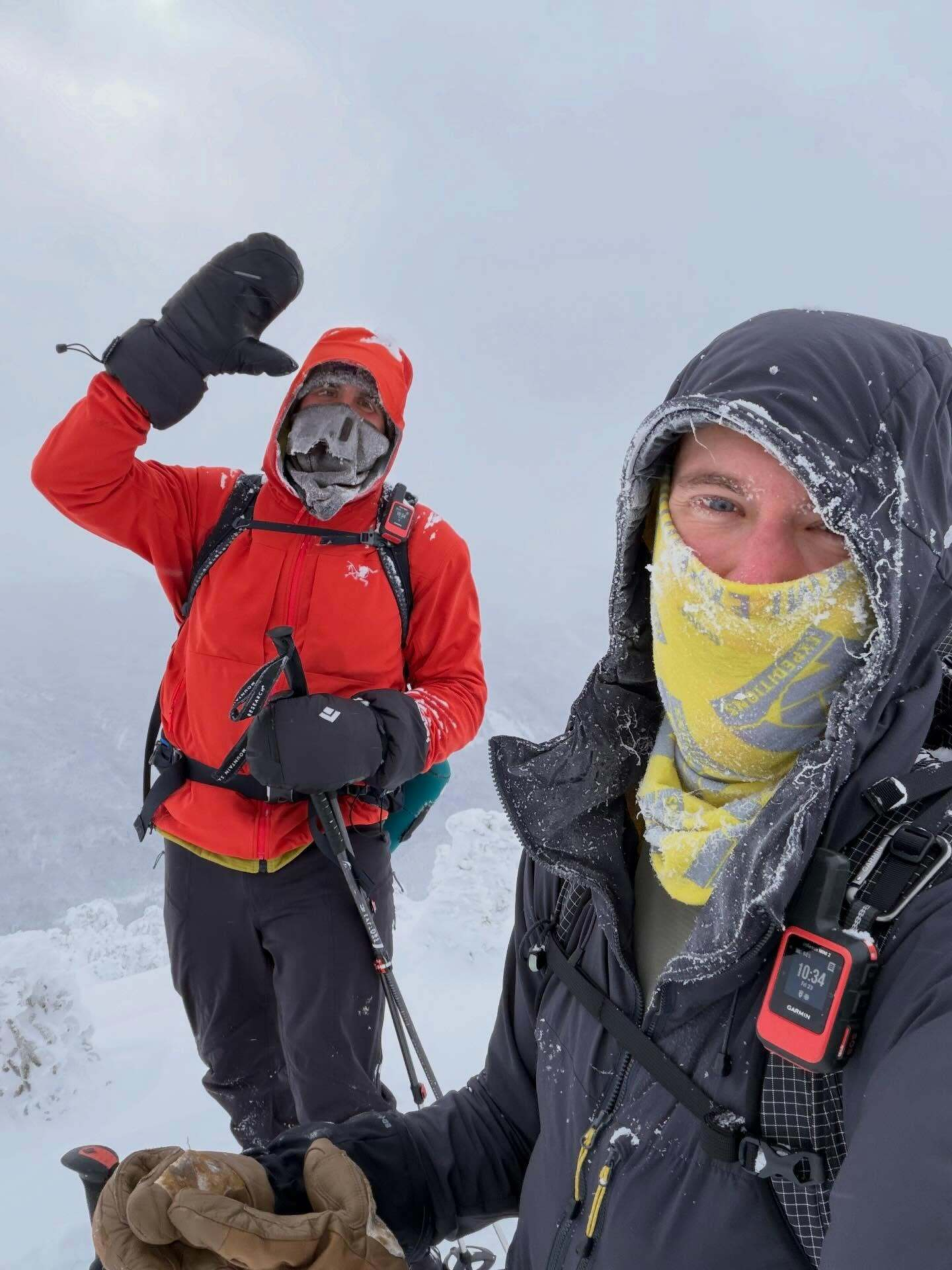 Two people stand on a snowy mountain peak wearing winter gear, including face coverings and gloves. One holds hiking poles, and the other displays a device showing 10 34 42 Sat.