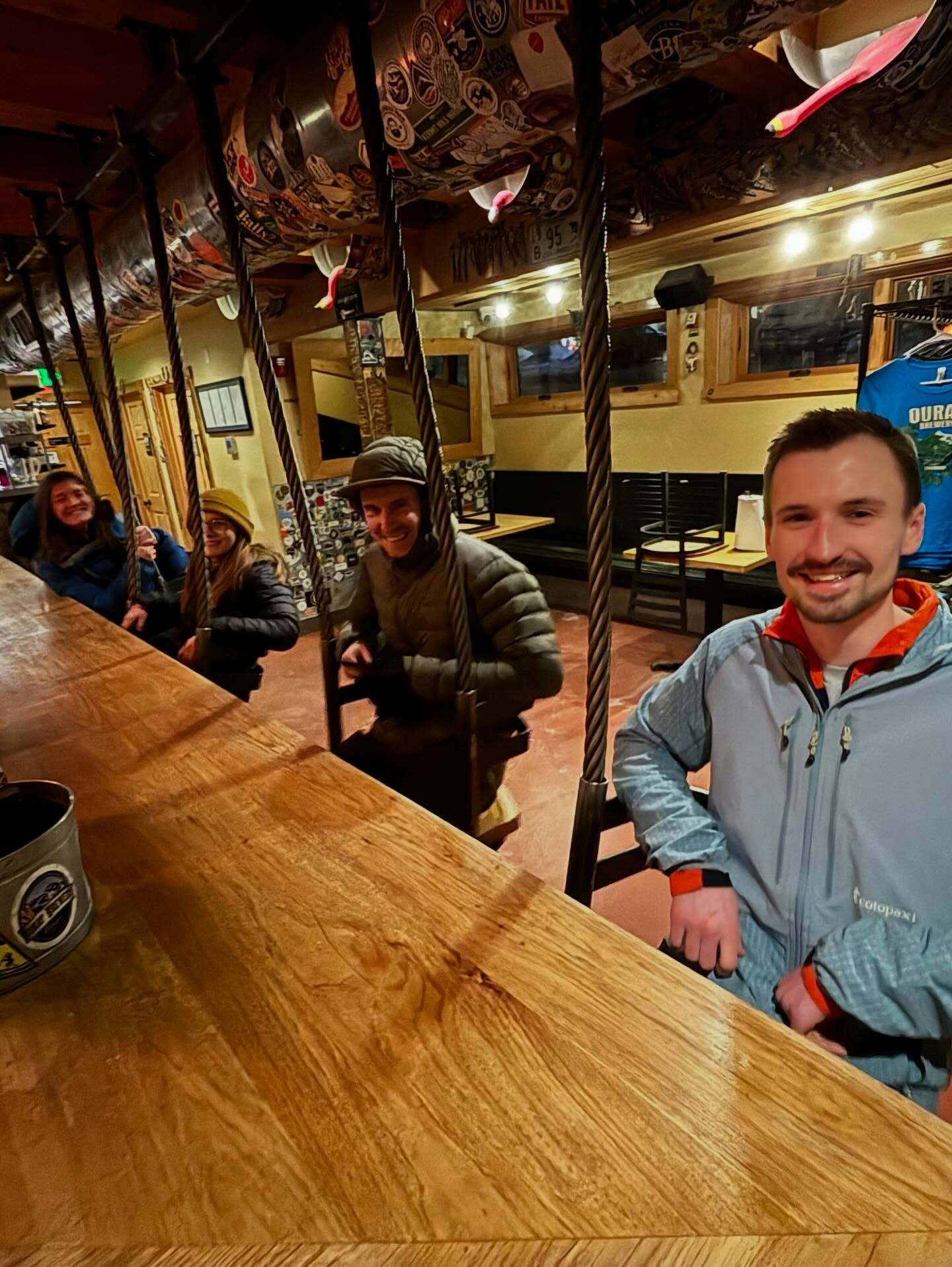 People sit on swing chairs attached to a wooden bar table. They smile and wear warm jackets. The setting is a cozy, sticker-decorated room with soft lighting and wooden furniture.