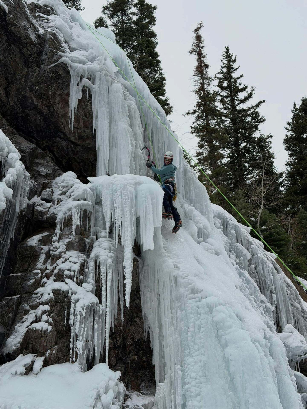 A climber ascends a steep icy cliff using ice axes surrounded by snow-covered rocks and evergreen trees under an overcast sky.