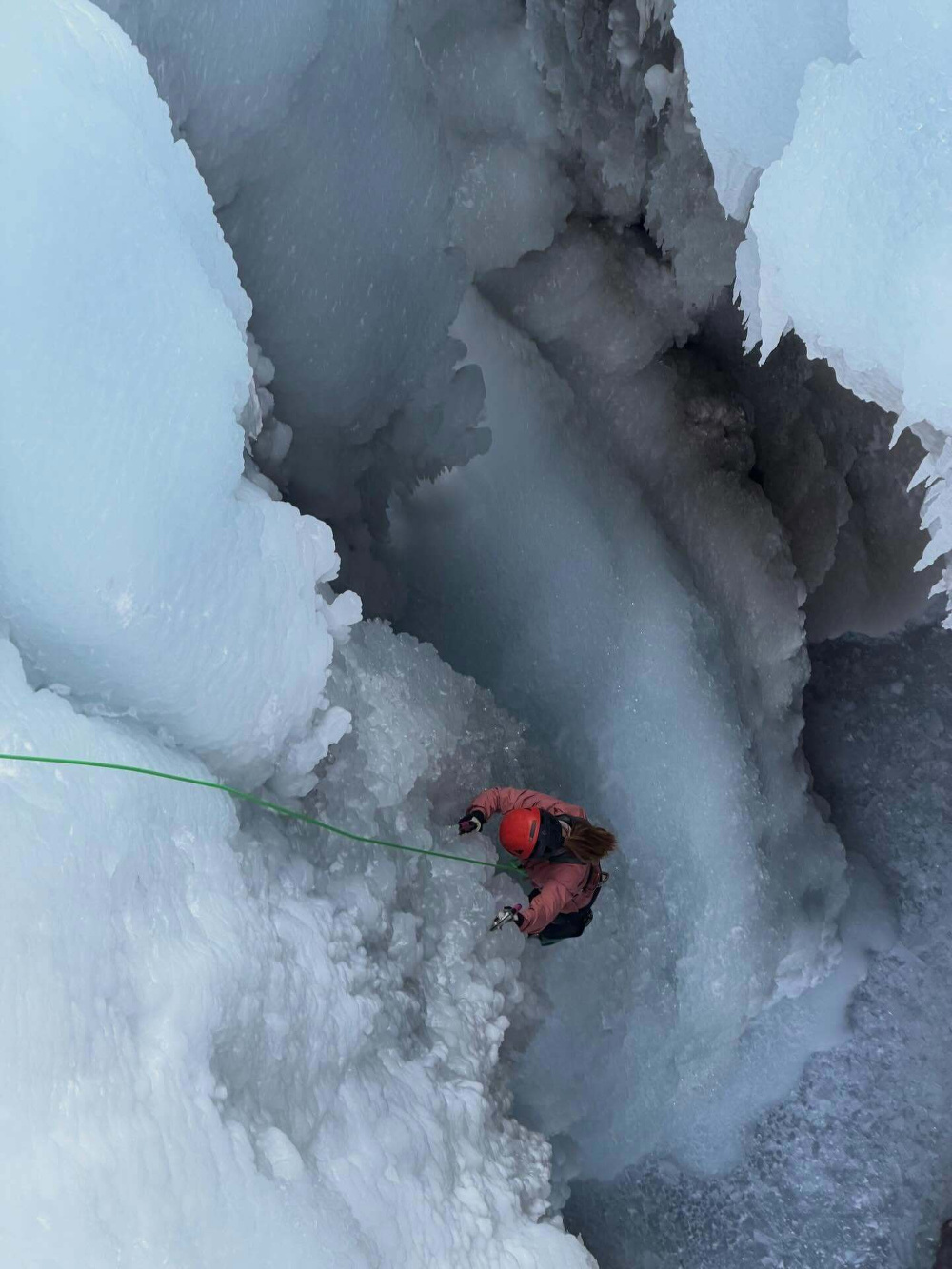 A person wearing a red helmet and climbing gear ascends an icy crevasse using a green rope surrounded by steep frozen walls in a challenging outdoor environment.