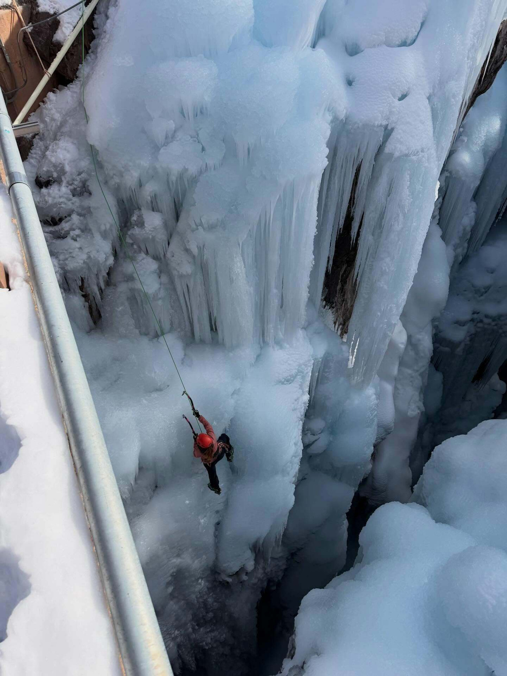 A climber ascends a frozen waterfall using ice axes and ropes surrounded by tall jagged ice formations with snowy ledges under a clear cold sky.