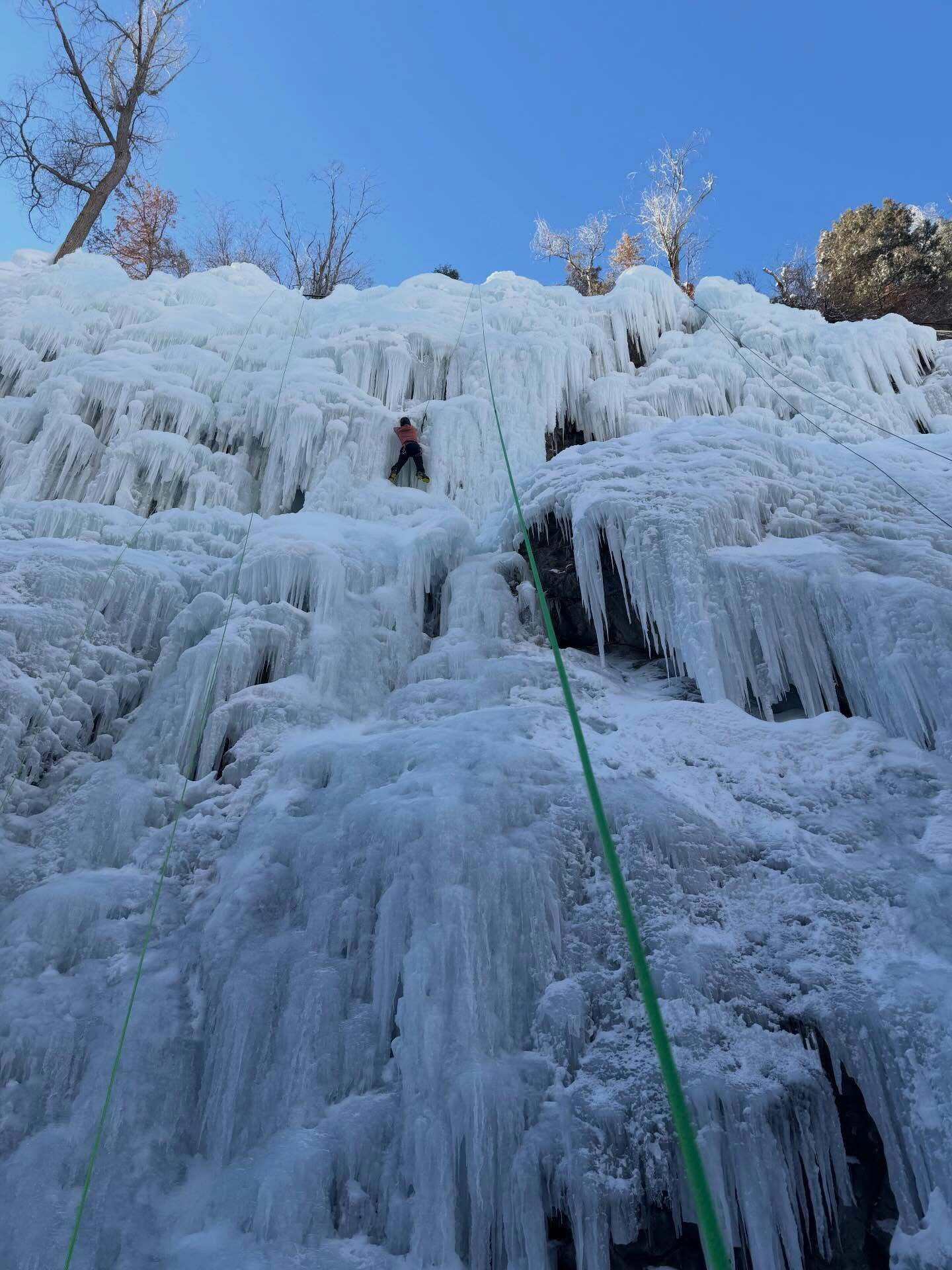 Climber ascends a frozen waterfall using an ice pick in a mountainous setting surrounded by ice formations and trees under a clear blue sky.