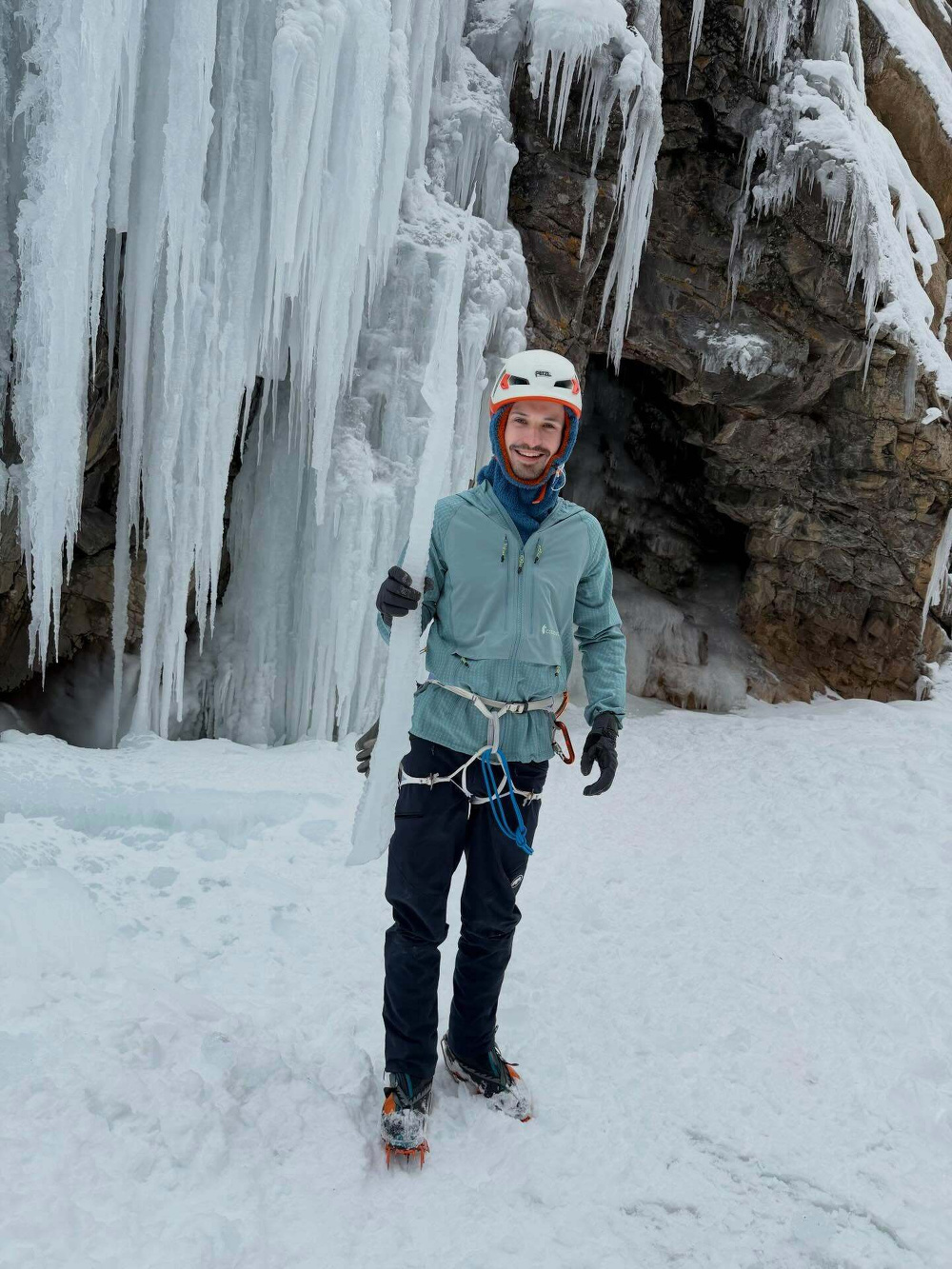 A person wearing climbing gear holds an icicle while standing on icy terrain. They are positioned near a wall of frozen icicles adjacent to a rocky cave entrance.