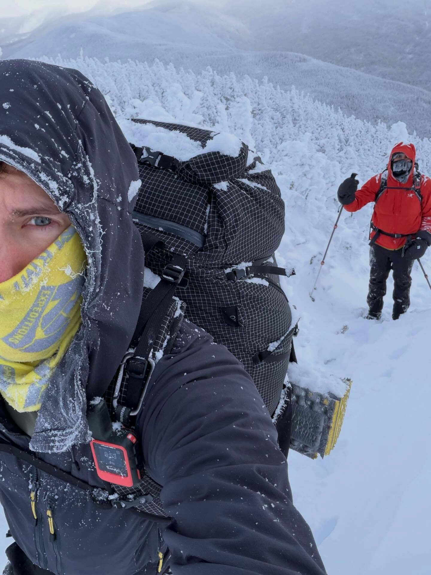 A hiker with a snow-covered backpack hikes up a snowy mountain trail while another person follows using trekking poles surrounded by a vast landscape of snow-laden trees and hills.