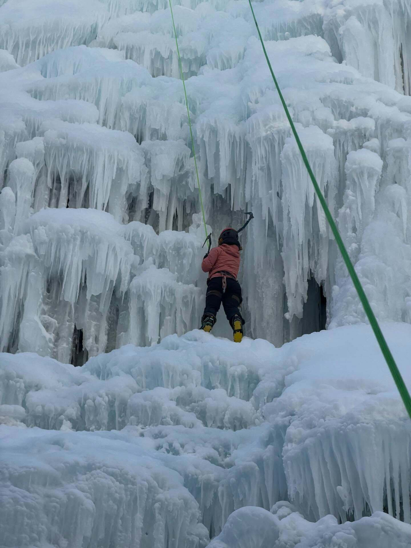 A person ascends an icy, frozen waterfall using climbing gear, surrounded by thick layers of ice forming irregular, jagged surfaces.