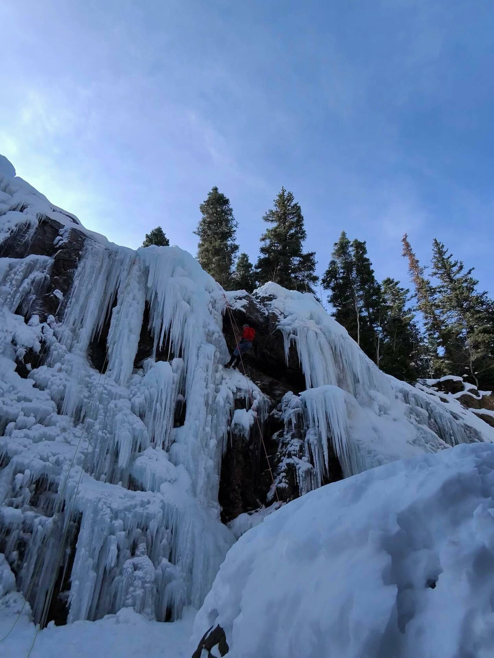 Climber ascends a frozen waterfall using ropes with pine trees visible at the top in a snowy mountain landscape under a clear blue sky.