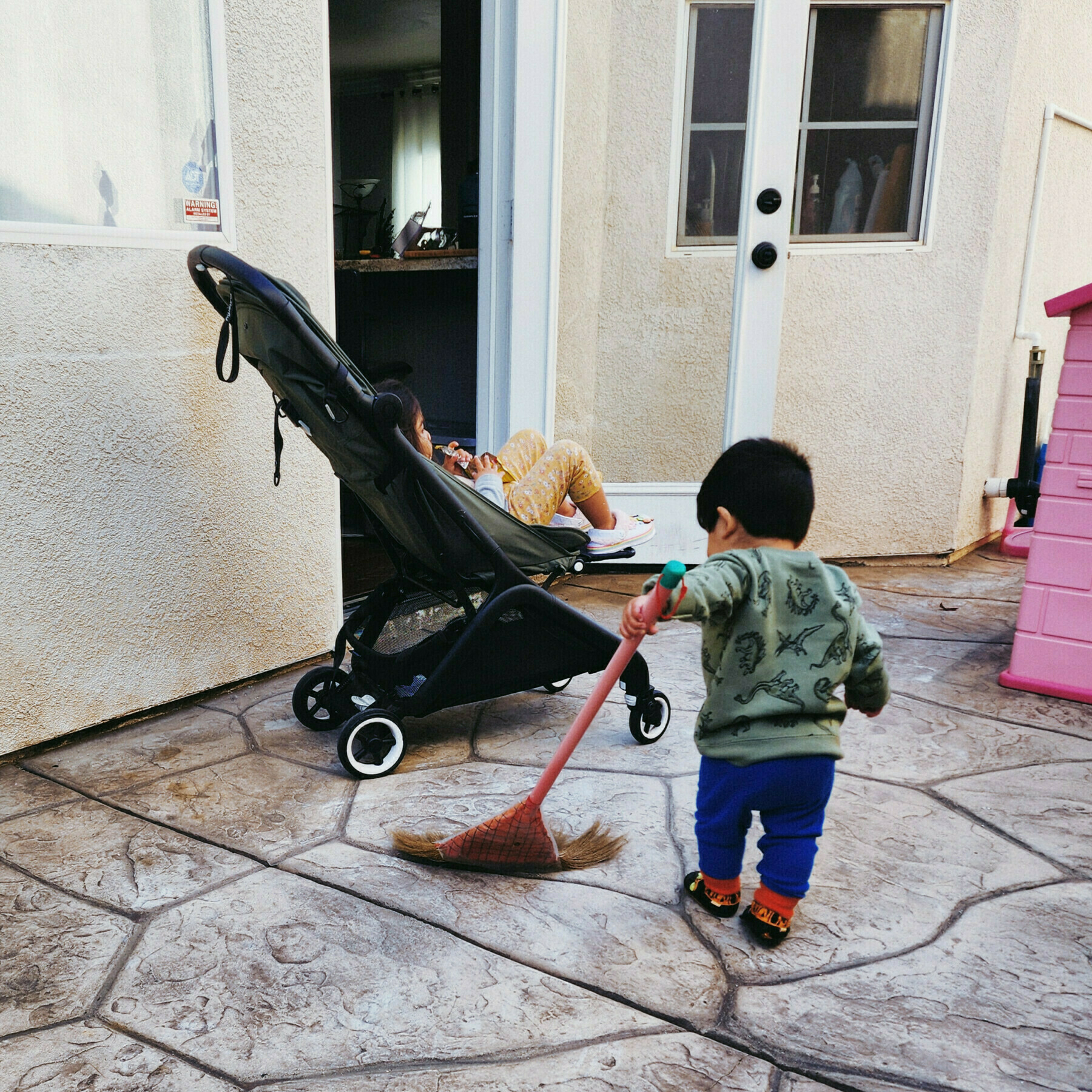 C— waiting in her stroller while J— sweeps the side yard with a broom his size
