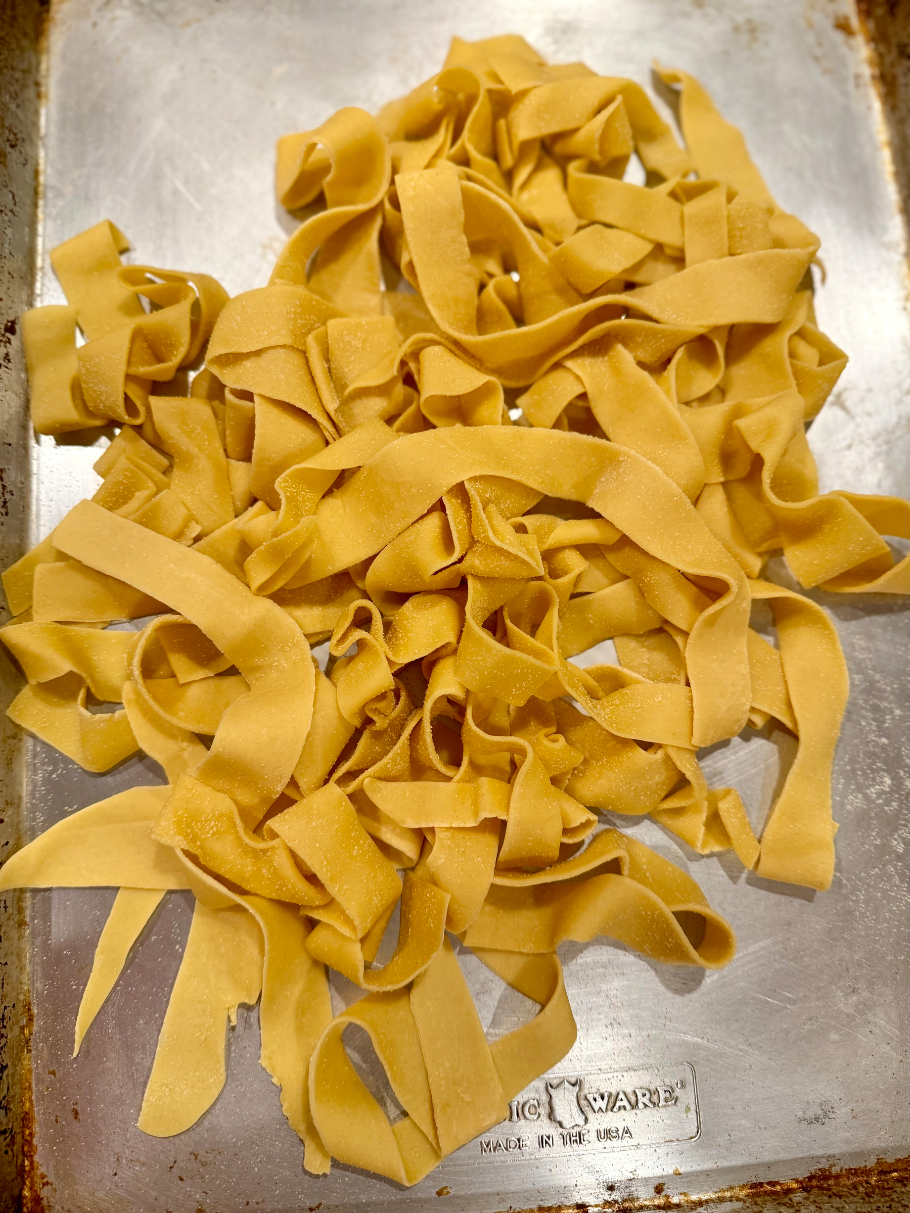 A pile of fresh, uncooked pasta ribbons is spread out on a metal baking tray.