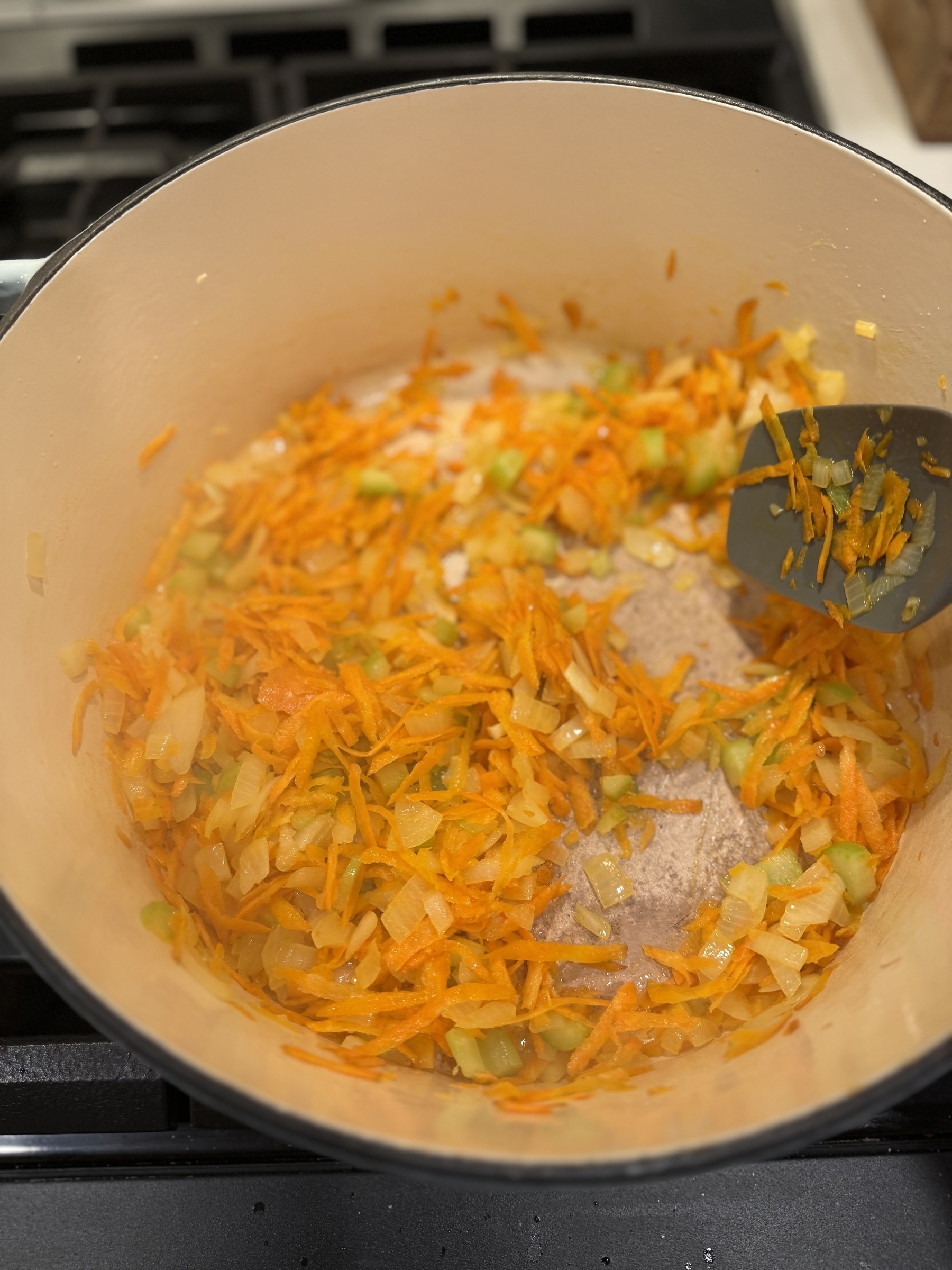 A pot contains a mixture of sautéed onions, carrots, and celery, being stirred with a spatula.