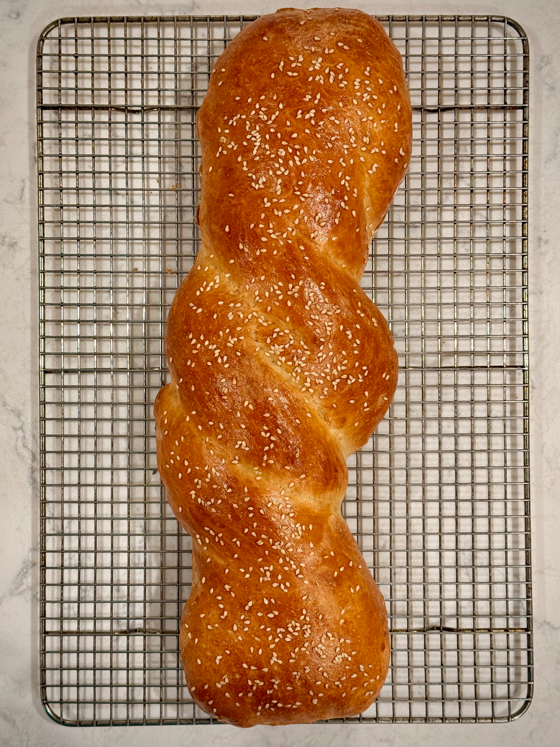 A freshly baked, golden-brown braided bread topped with sesame seeds rests on a cooling rack.