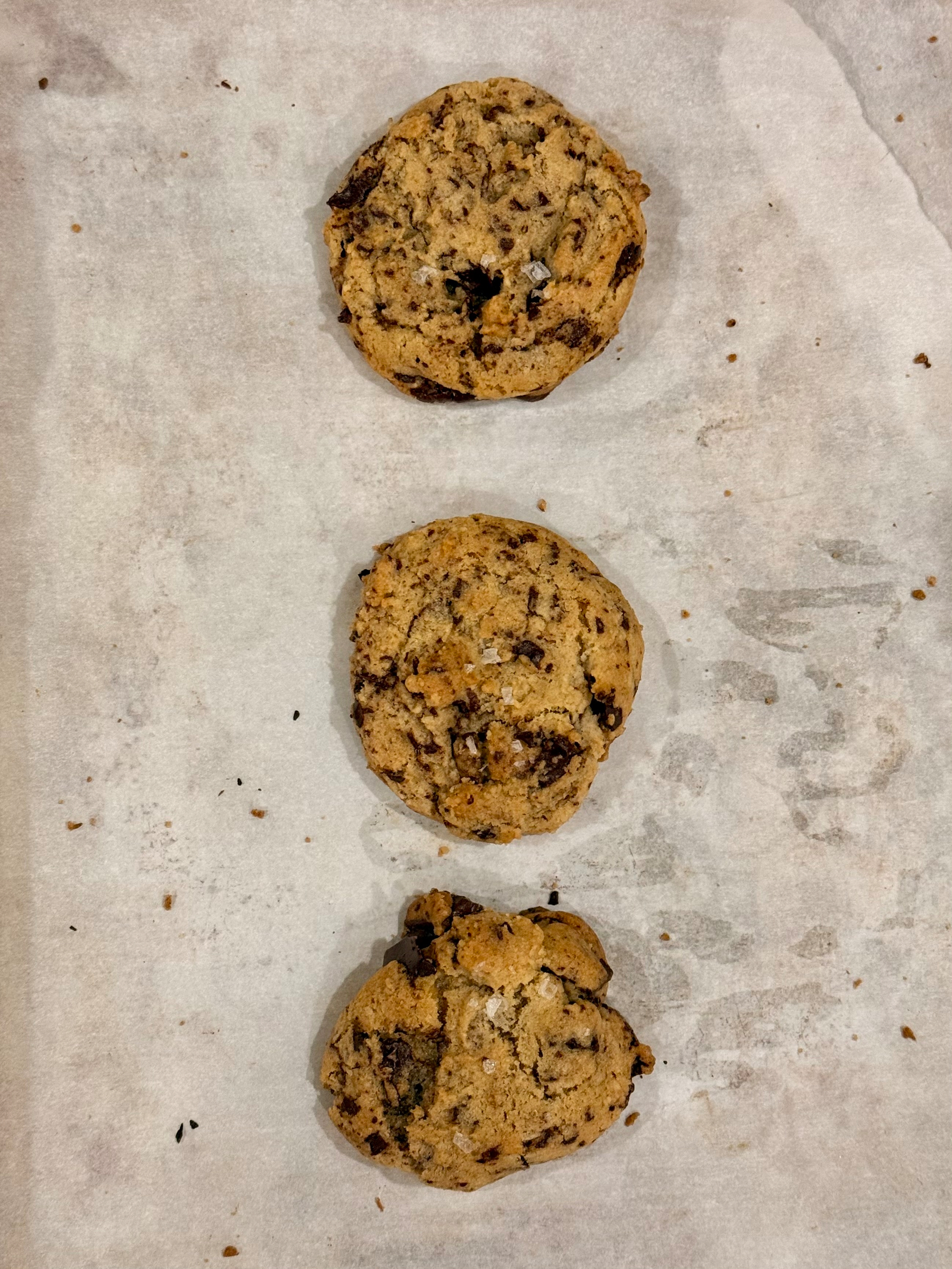 Three chocolate chip cookies are arranged vertically on a sheet of parchment paper.