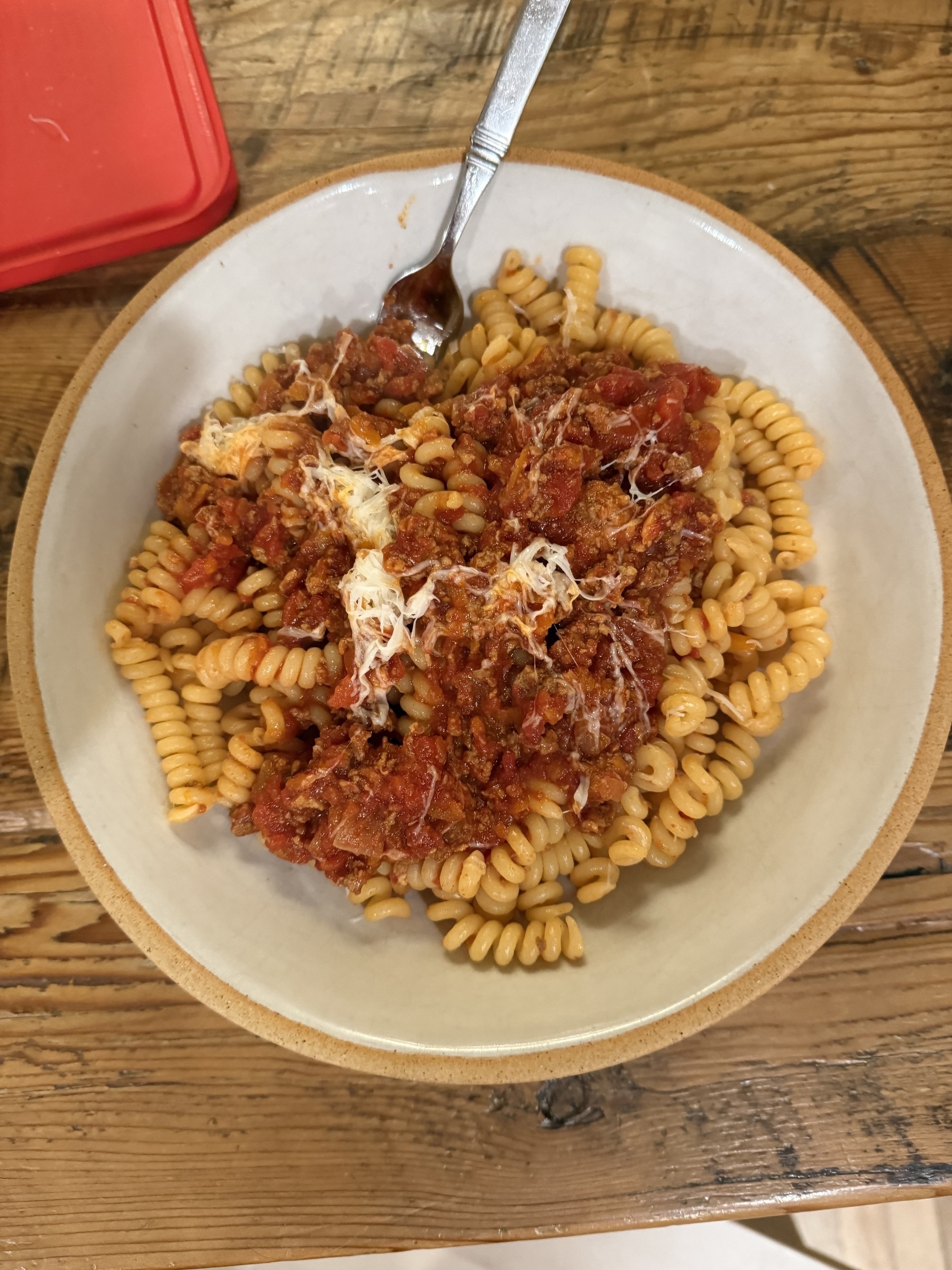 A bowl of corkscrew pasta topped with a chunky red sauce and shredded cheese is placed on a wooden surface.
