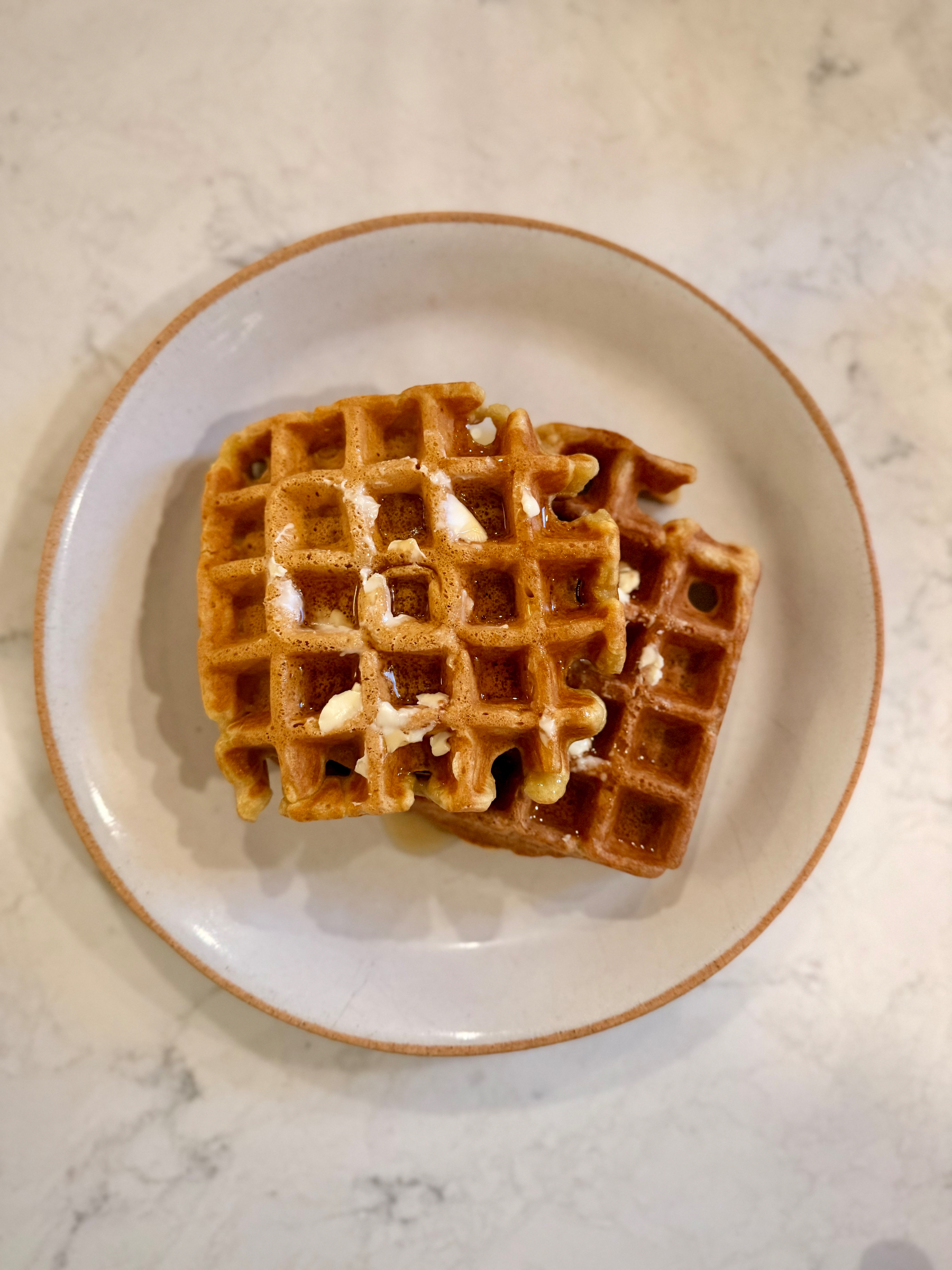 Two waffles with butter are served on a white plate against a marble background.