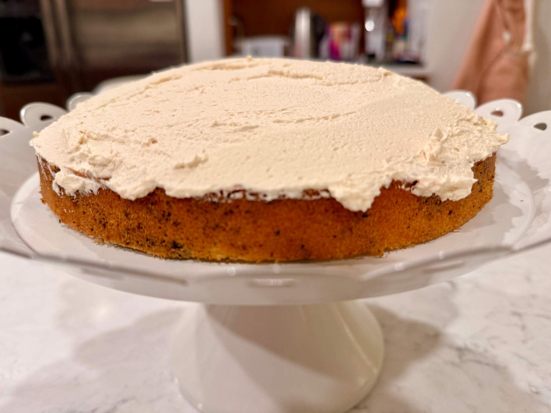 A cake with light-colored frosting sits on a white cake stand.