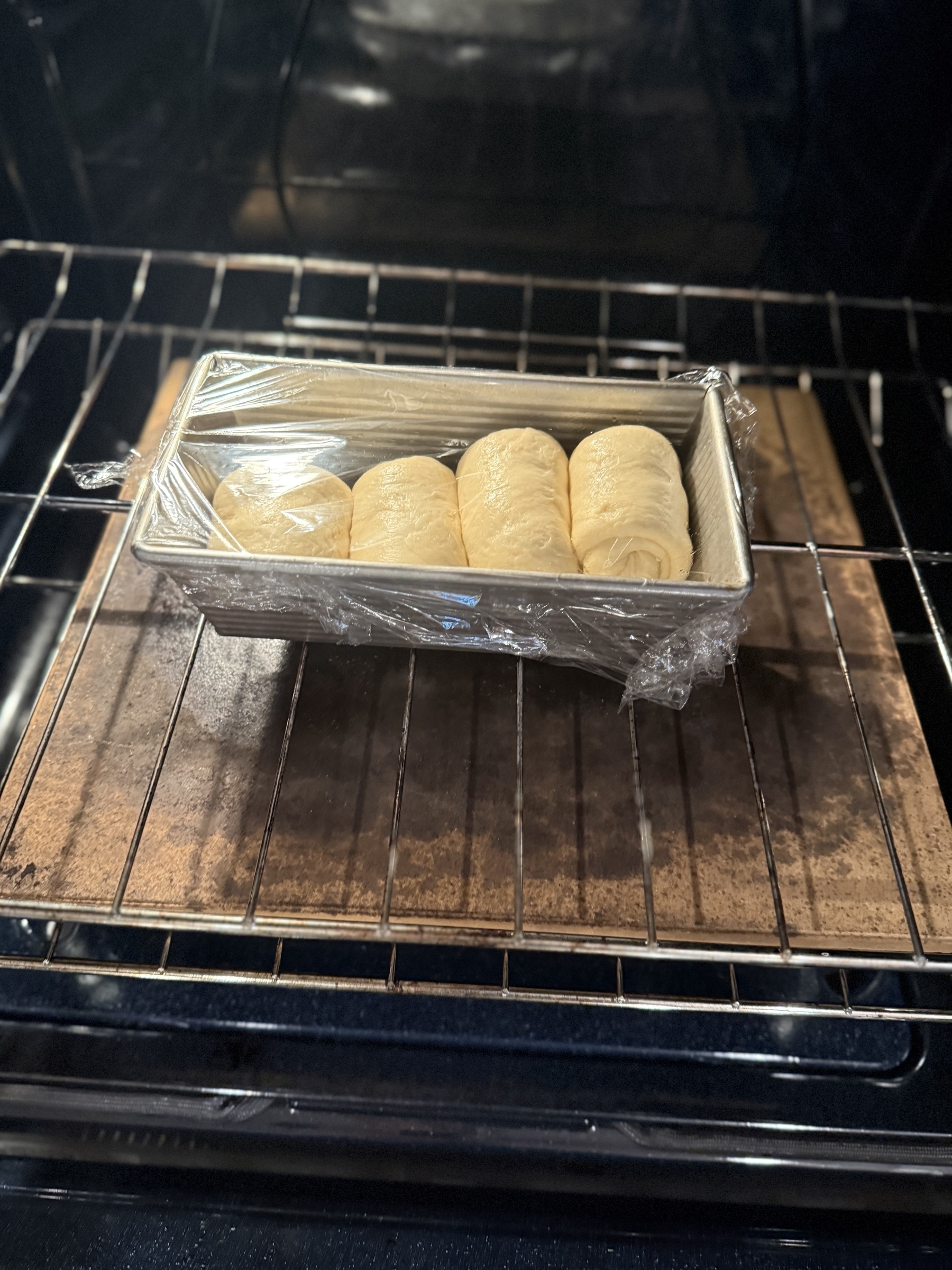 A loaf pan with rising dough rolls covered in plastic wrap is placed in an oven.