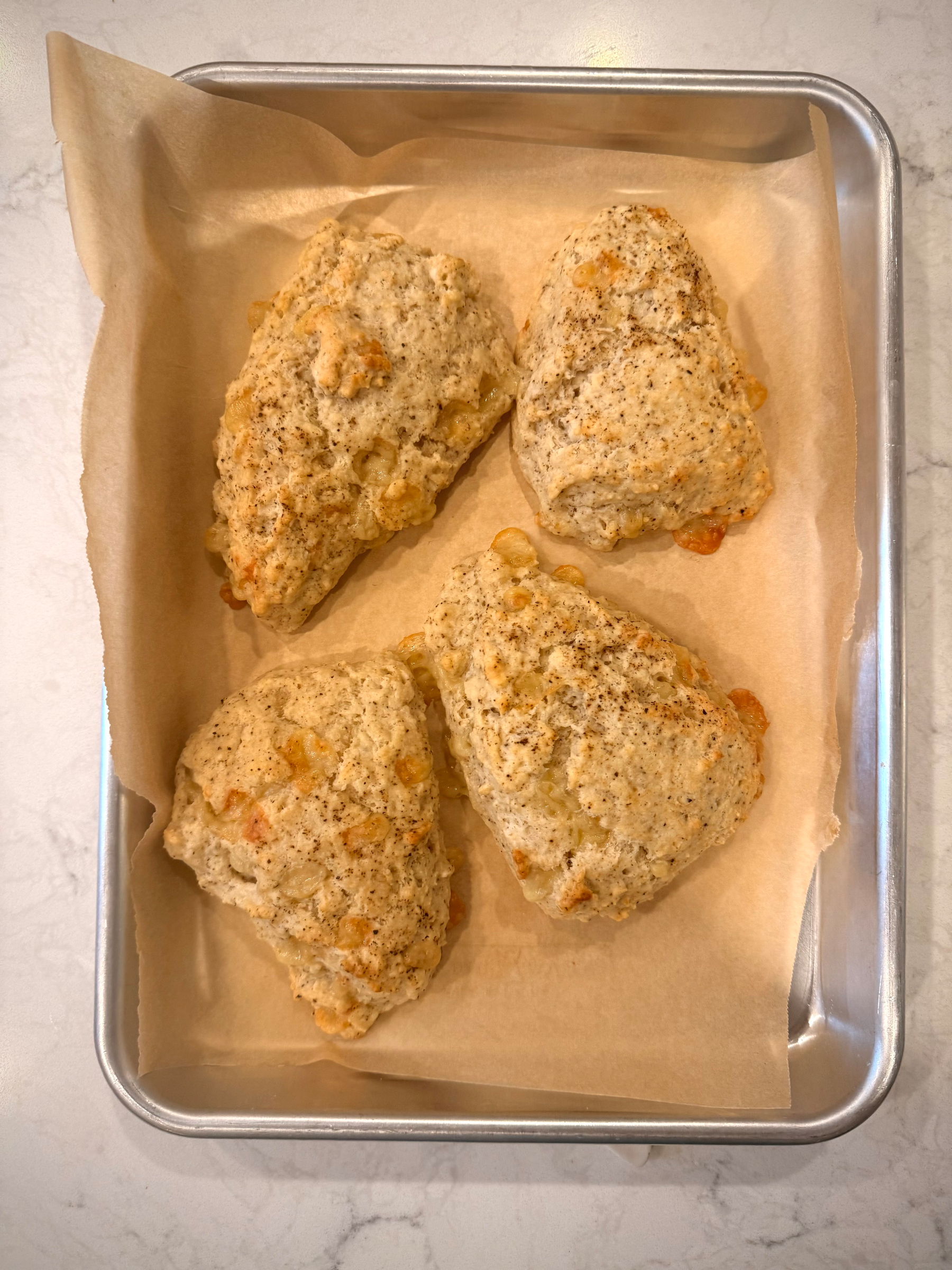 Four baked scones with a golden, slightly craggy surface sit on a parchment-lined sheet pan, showing bits of melted cheese or mix-ins on top.
