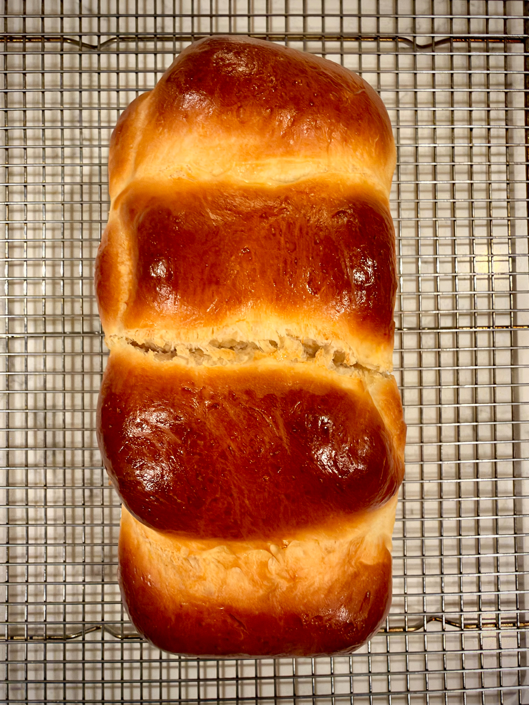 A freshly baked loaf of bread with a shiny golden-brown crust rests on a metal cooling rack.