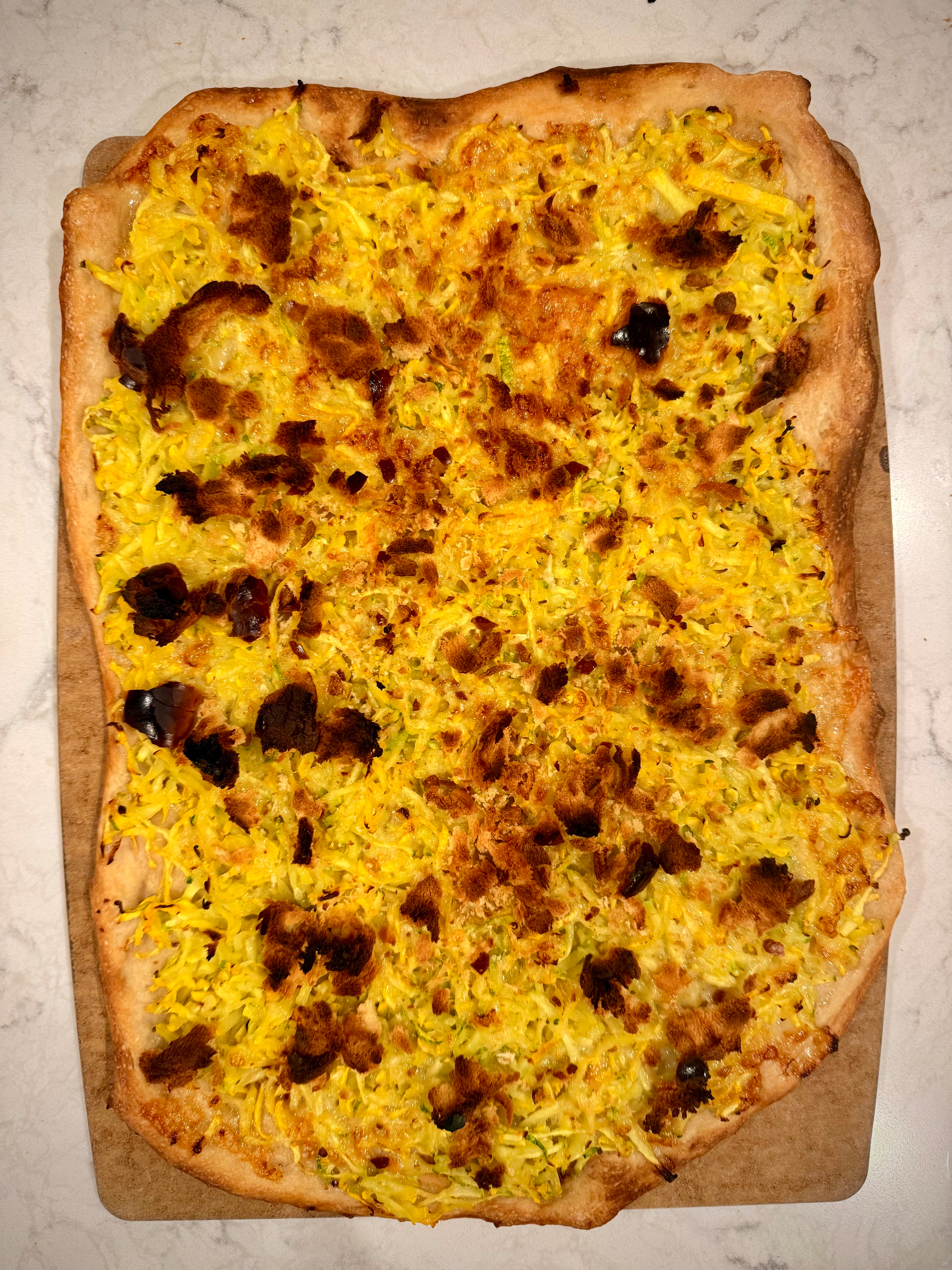 A rectangular pizza with a crispy crust is topped with melted cheese, shredded squash and scattered browned spots.