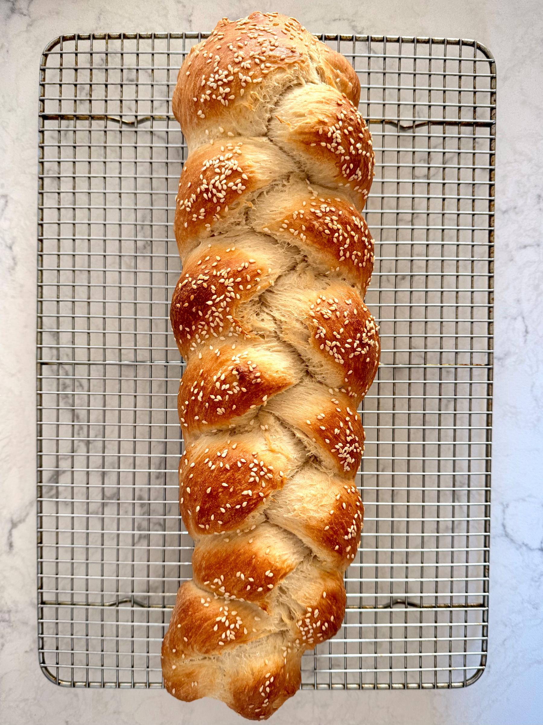 A freshly baked, braided loaf of bread topped with sesame seeds rests on a cooling rack.