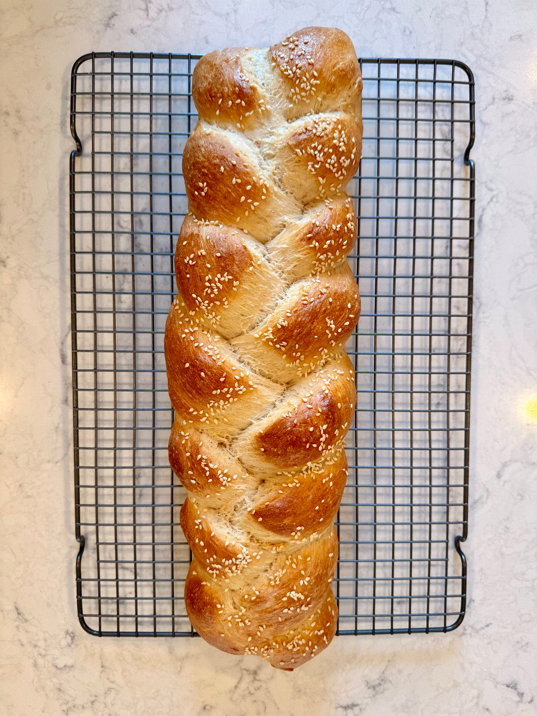 A freshly baked braided loaf of bread with sesame seeds is resting on a cooling rack.