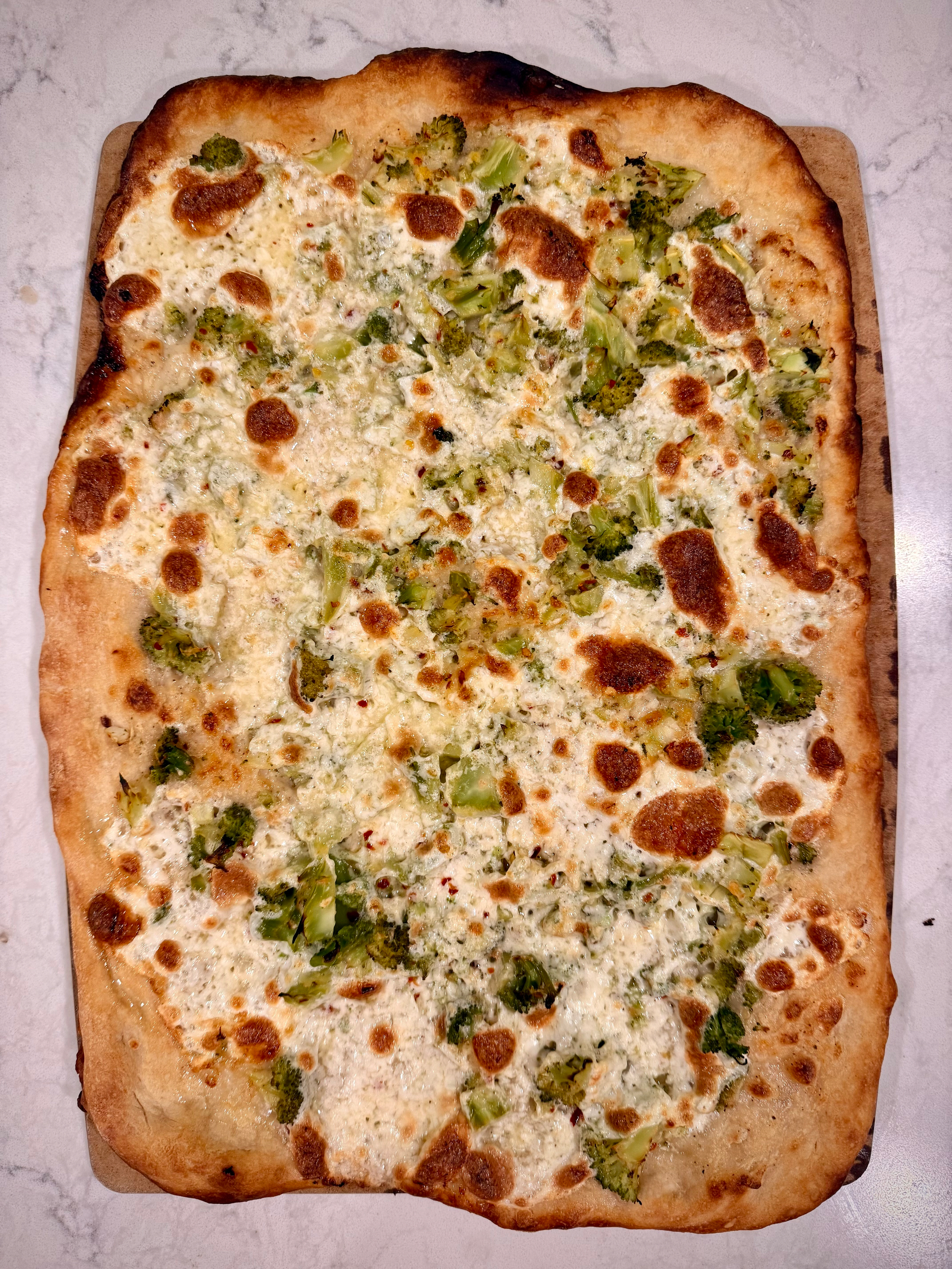A rectangular pizza topped with cheese and pieces of broccoli is placed on a wooden board.